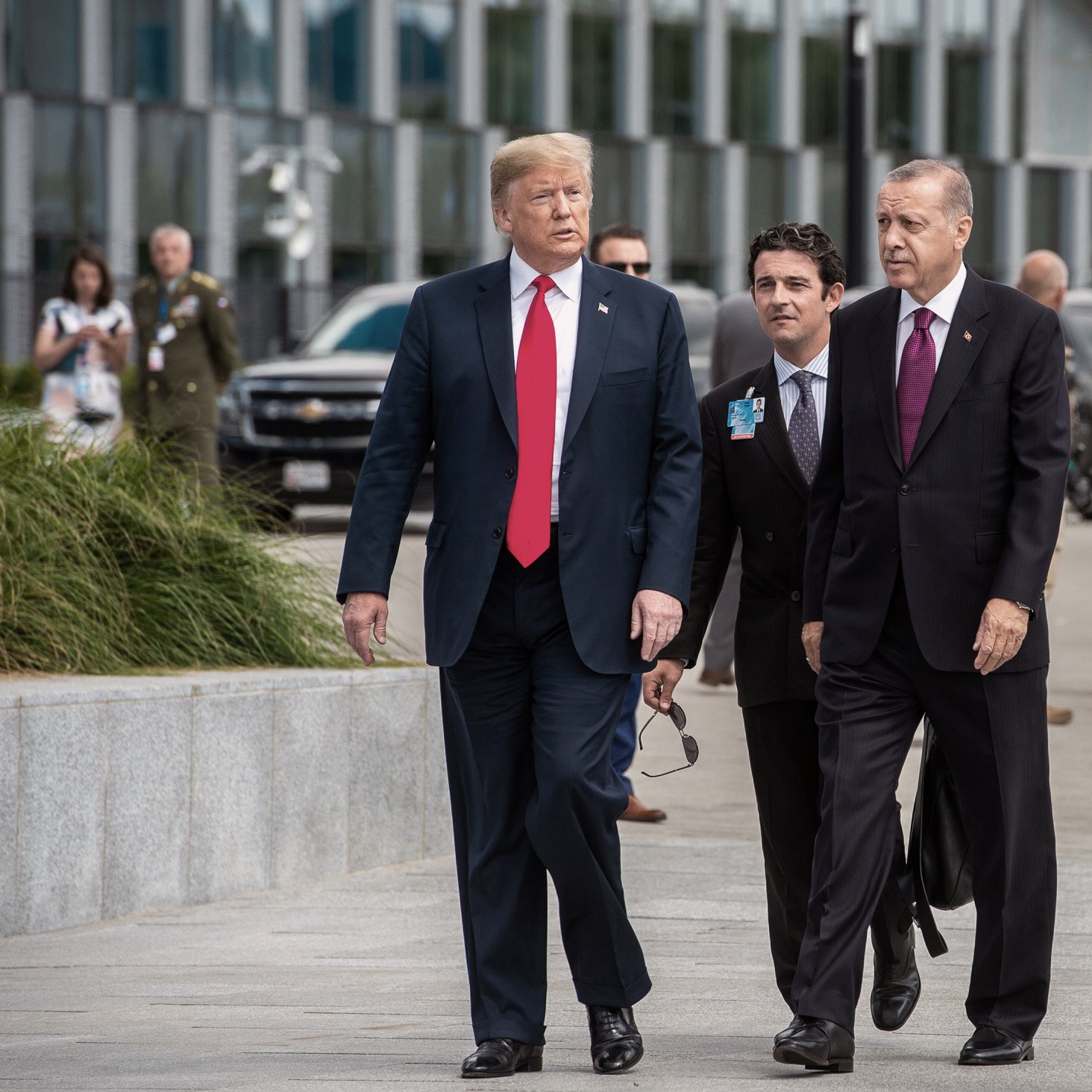 در این تصویر، چند مرد در حال پیاده‌روی هستند. یکی از آنها که لباس رسمی پوشیده و کلاهی تیره به سر دارد، به سمت دیگران نگاه می‌کند. شخص دیگری که در سمت چپ او قرار دارد، کت و شلوار و یک کراوات قرمز به تن دارد. دو مرد دیگر نیز به آنها ملحق شده‌اند و ظاهراً در حال گفتگو هستند. پس‌زمینه، شامل ساختمان‌های مدرن و تعدادی از افراد دیگر در حال حرکت است. جو کلی تصویر نشان‌دهنده یک ملاقات رسمی است.