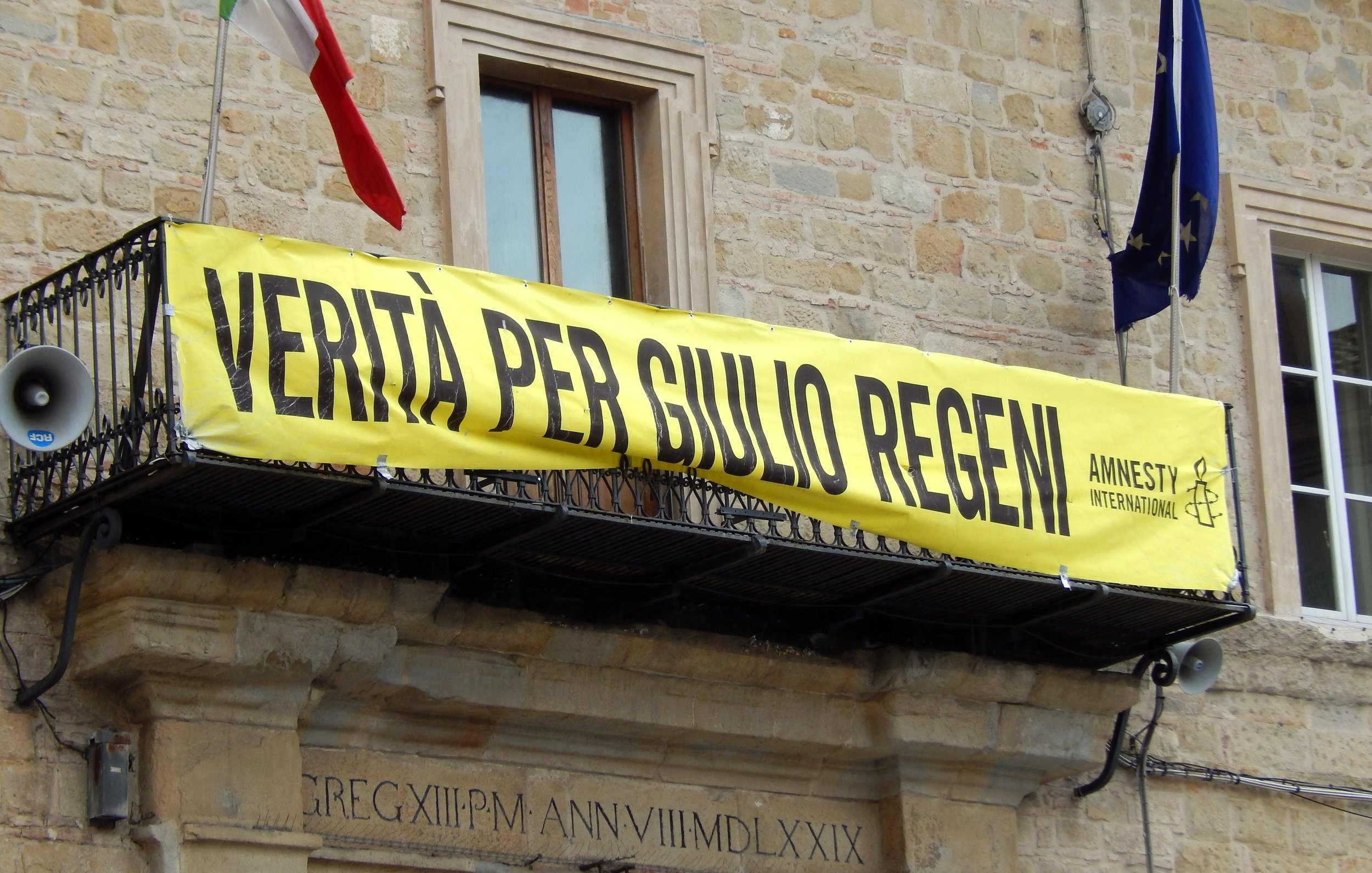 L'immagine mostra un grande striscione giallo appeso a un balcone di un edificio. Sullos striscione è scritto "VERITÀ PER GIULIO REGENI", accompagnato dal logo di Amnesty International. Nel contesto, si possono notare delle bandiere, probabilmente italiane, che sventolano. L'atmosfera suggerisce una manifestazione di sostegno e richiesta di giustizia per Giulio Regeni, un caso di rilevanza pubblica.