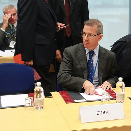 L'image montre une scène de réunion où plusieurs personnes sont présentes autour d'une table. Un homme, assis au premier plan, porte un costume gris et une cravate bleue à pois. Il semble concentré, en train d'organiser des documents devant lui. Sur la table, il y a des bouteilles d'eau et des documents disposés. À l'arrière-plan, d'autres participants sont visibles, certains discutant ou se déplaçant. L'environnement semble formel, typique d'une conférence ou d'une réunion professionnelle.