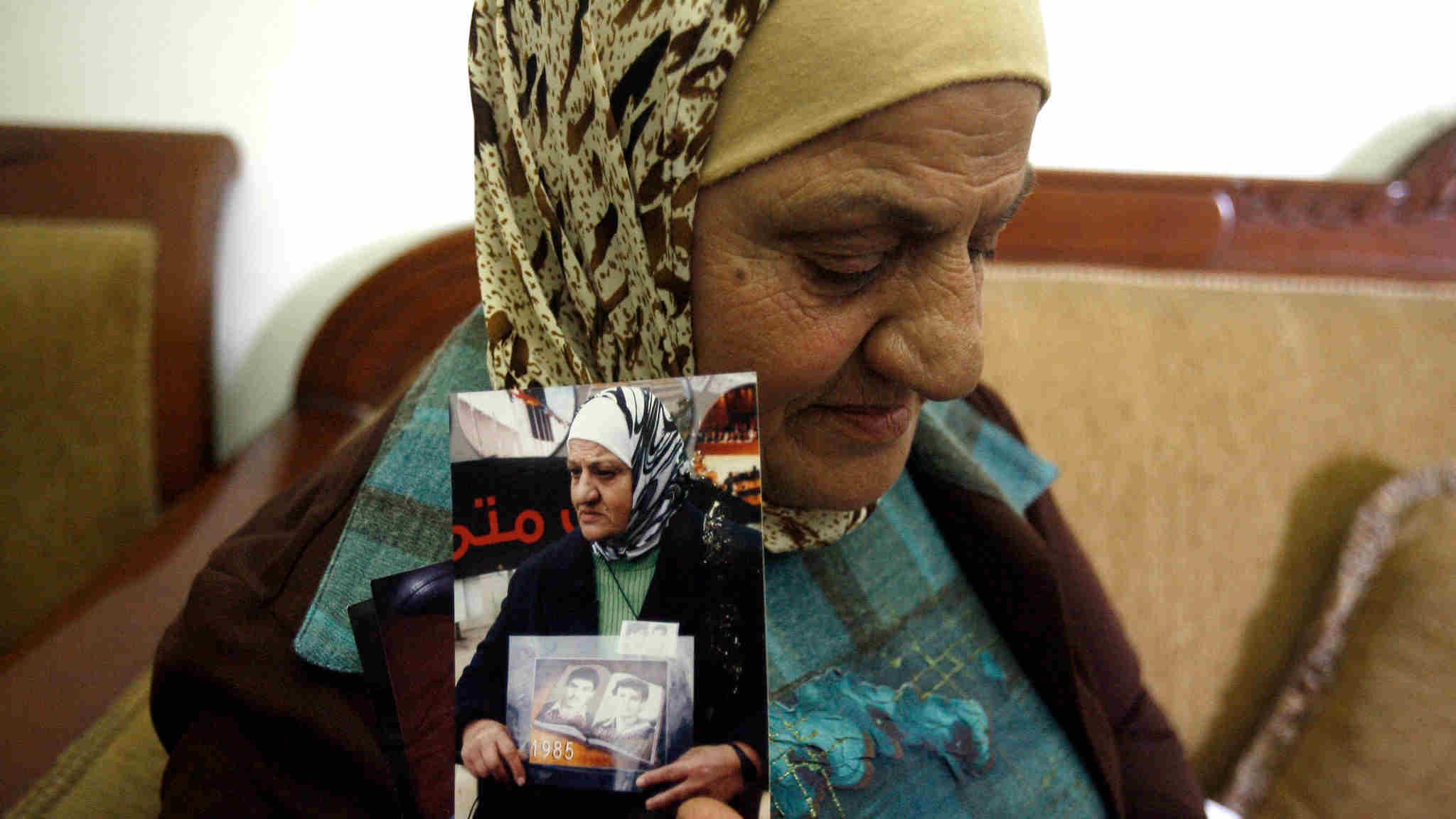 الصورة تظهر سيدة كبيرة في السن ترتدي حجابًا وتجلس على أريكة. تعبر ملامح وجهها عن التأمل والحنين، حيث تُمسك بعدد من الصور الفوتوغرافية في يديها. يبدو أن هذه الصور تحمل ذكريات مهمة بالنسبة لها، وقد تكون تتعلق بأفراد من عائلتها. الخلفية بسيطة وتُشير إلى بيئة منزلية مريحة.