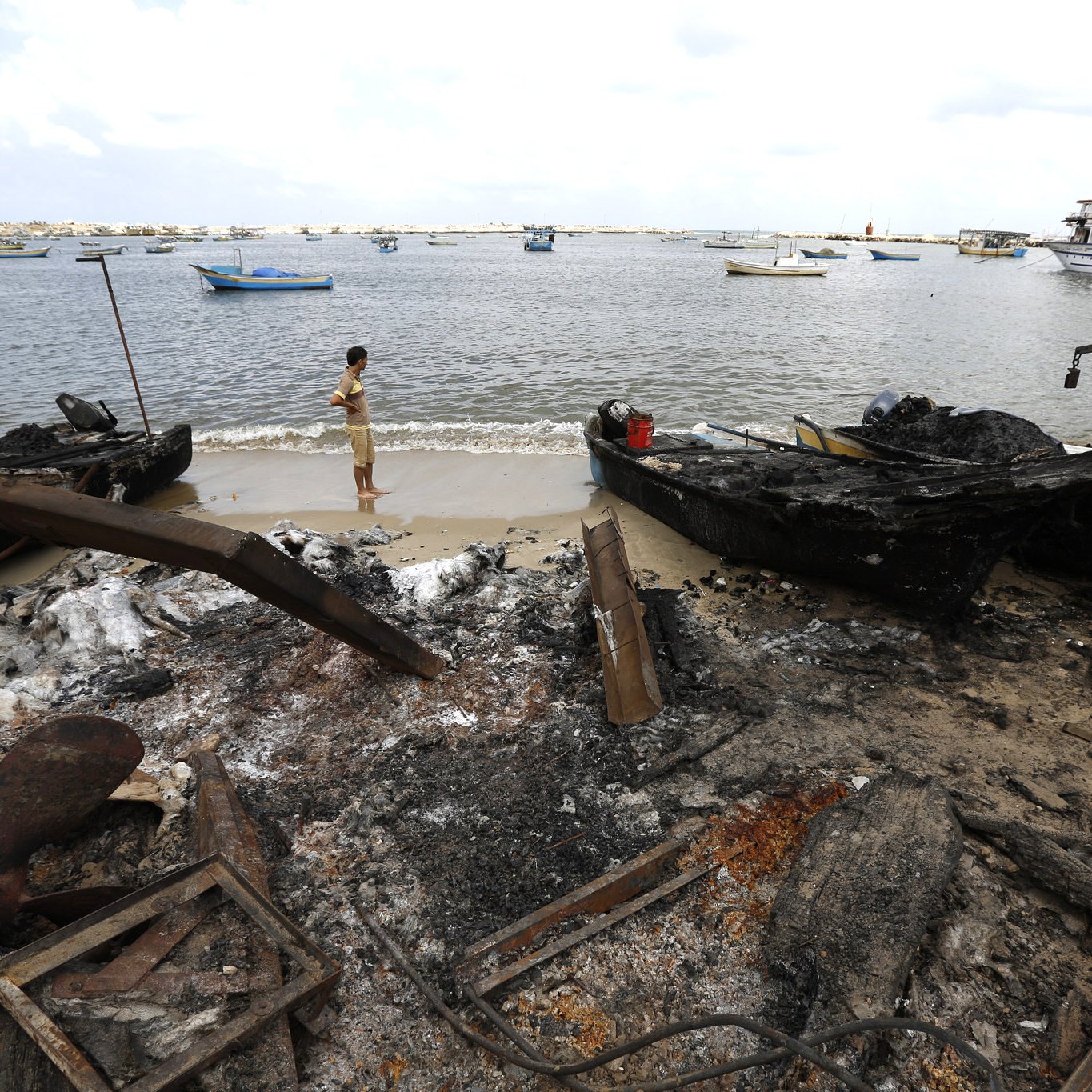 L'image montre une scène côtière après un incendie. On peut y voir des bateaux endommagés et carbonisés sur le rivage. Un homme se tient près de l'eau, regardant vers le horizon. La mer semble calme, avec plusieurs petites embarcations au loin. Le ciel est nuageux, et le paysage est marqué par les conséquences de l'incendie, avec des débris et des cendres sur le sable.