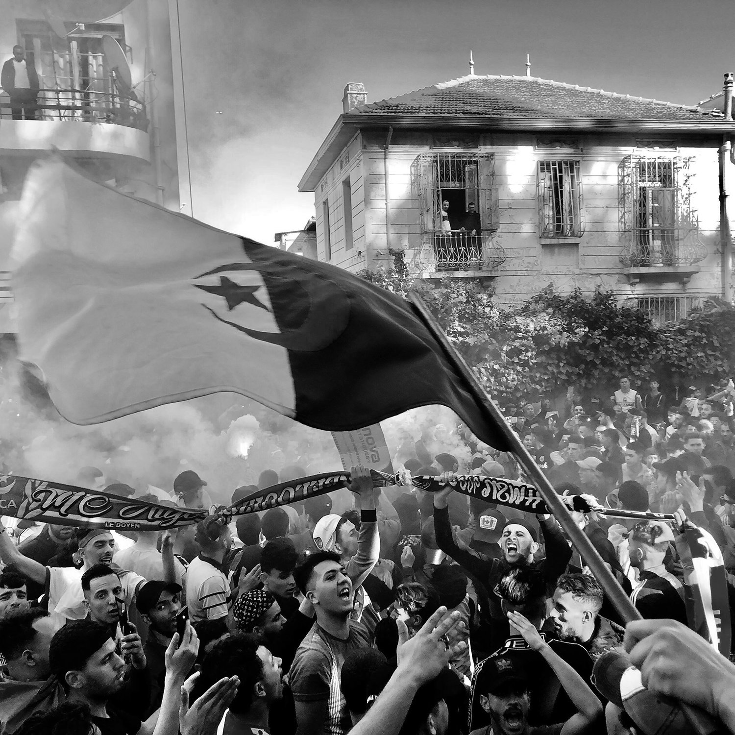 La imagen muestra a una gran multitud en un ambiente festivo, probablemente durante un evento deportivo o una celebración. Se pueden ver personas ondeando una bandera, así como pancartas y bufandas, lo que sugiere un fuerte sentido de identidad y apoyo a un equipo. El ambiente está lleno de humo, lo que añade un efecto dramático a la escena. La gente parece emocionada y unida, disfrutando del momento. Las edificaciones al fondo indican que la actividad está ocurriendo en un área urbana.
