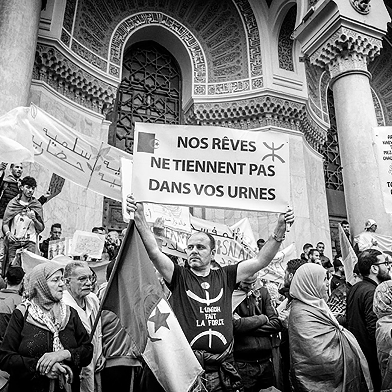 The image depicts a protest scene, where a diverse group of people is gathered. Central to the scene is a man holding a large sign that reads "NOS RÊVES NE TIENNENT PAS DANS VOS URNES," which translates to "Our dreams do not fit in your ballot boxes." The crowd appears to be passionate, with some individuals holding banners, possibly conveying political messages. The setting has an architectural backdrop, suggesting a significant public space, such as a government building or place of gathering. The black and white tones emphasize the seriousness of the demonstration.
