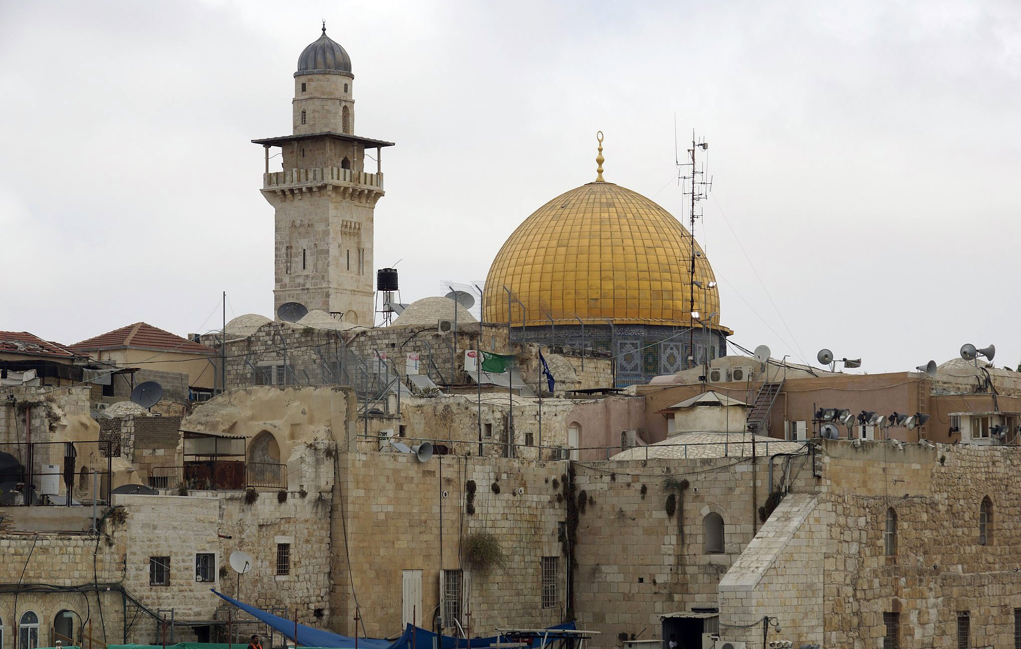 تظهر الصورة مشهداً لمدينة القدس، حيث يتوسطها المسجد الأقصى، الذي يبرز بقبة ذهبية مميزة. بجانب القبة، يمكن رؤية مئذنة ترتفع عالياً، وهناك بعض المباني التاريخية ذات الحجر القديم. كما يظهر في الصورة وجود أجهزة استقبال الأقمار الصناعية على أسطح المباني، مما يعكس مزيجاً من التاريخ والحداثة في هذه المنطقة. السماء تبدو غائمة، مما يضفي على المشهد جوّاً من السكون.
