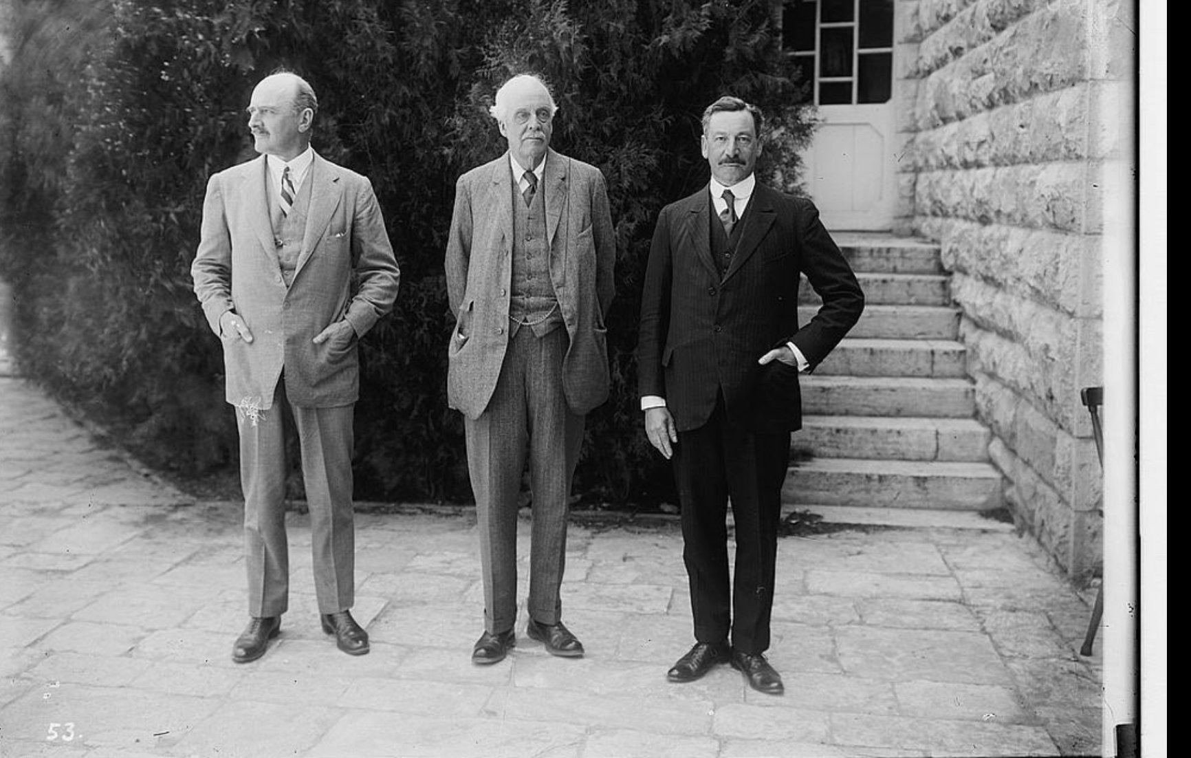 سه مرد در لباس رسمی ایستاده‌اند. پس‌زمینه شامل درختان و یک سازه سنگی ساده است.