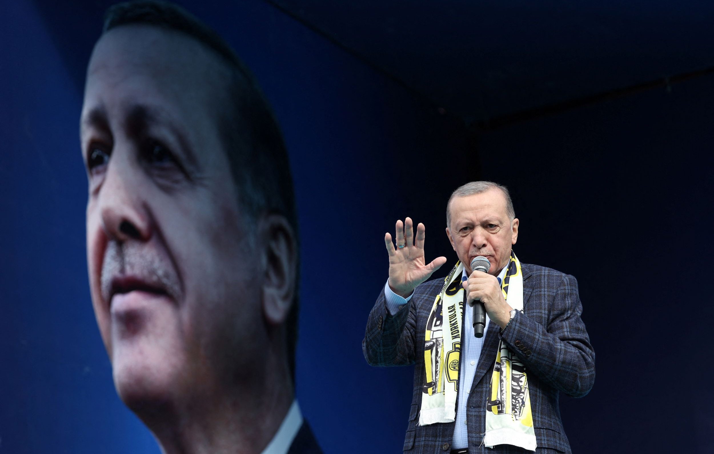 Nell'immagine si vede un uomo che parla a un pubblico, indossando una giacca a quadri e una sciarpa. Dietro di lui c'è un grande murale o un'immagine di un volto che lo ritrae. L'ambientazione sembra essere un evento pubblico o un comizio, con un'atmosfera di fervore e partecipazione. Il fondo è di colore blu, creando un contrasto con il soggetto principale.