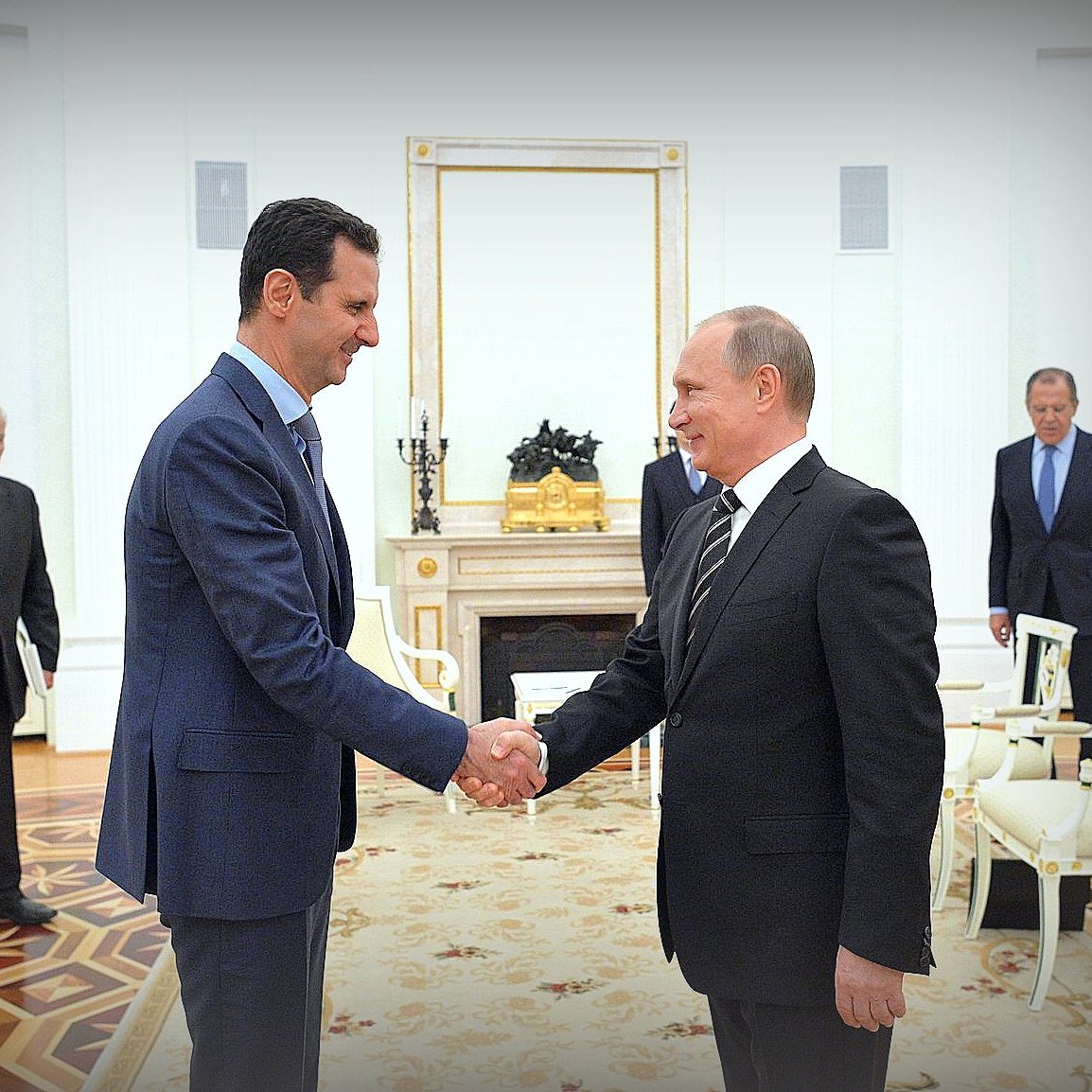 The image depicts a meeting between two men shaking hands in an ornate room. One person appears to be dressed in a suit and has a distinguished demeanor, while the other is also in formal attire. They are surrounded by a few other individuals in the background, all dressed formally. The setting has an elegant decor with artwork on the walls and a polished floor, suggesting a formal diplomatic meeting or discussion.