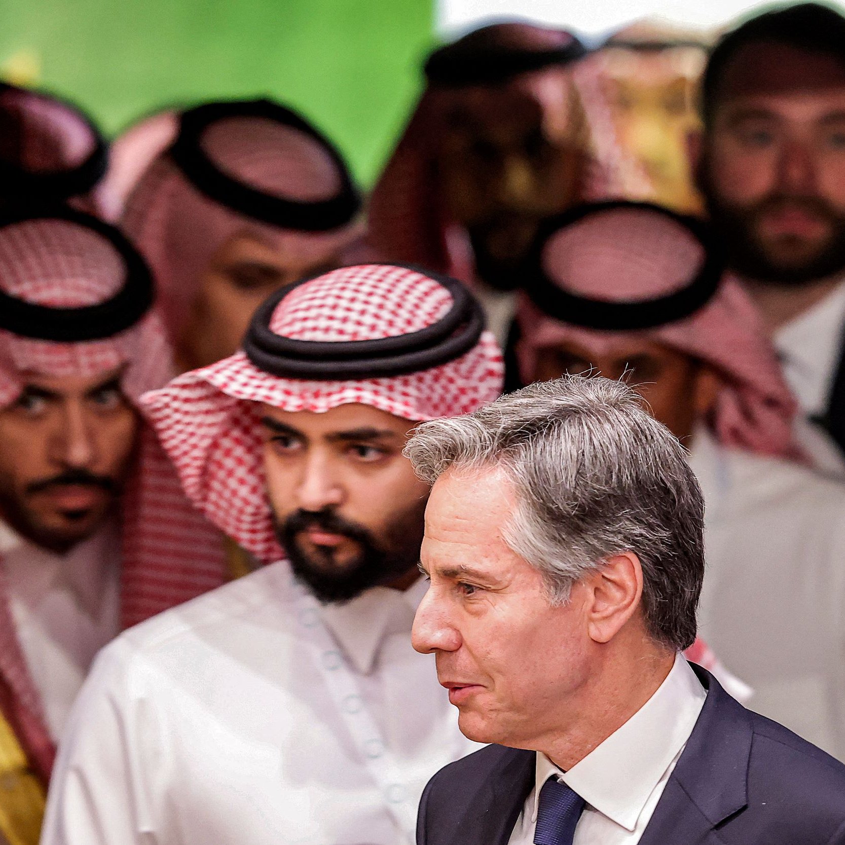 L'image montre un groupe d'hommes en vêtements traditionnels saoudiens, portant des ghoutas et des agals, en arrière-plan. Au premier plan, un homme en costume sombre marche, semblant engagé dans une conversation ou une réflexion. L'ambiance générale de l'image suggère une réunion ou un événement diplomatique.