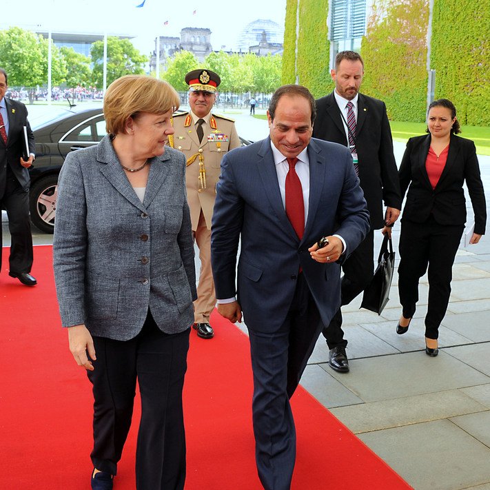 L'image montre un moment officiel, probablement lors d'une rencontre diplomatique. Une femme, portant un blazer gris, marche sur un tapis rouge aux côtés d'un homme en costume sombre. Ils semblent engagés dans une conversation. À l'arrière-plan, on peut voir des personnes en costume, ainsi que des bâtiments modernes et une végétation. L'atmosphère est formelle et professionnelle.