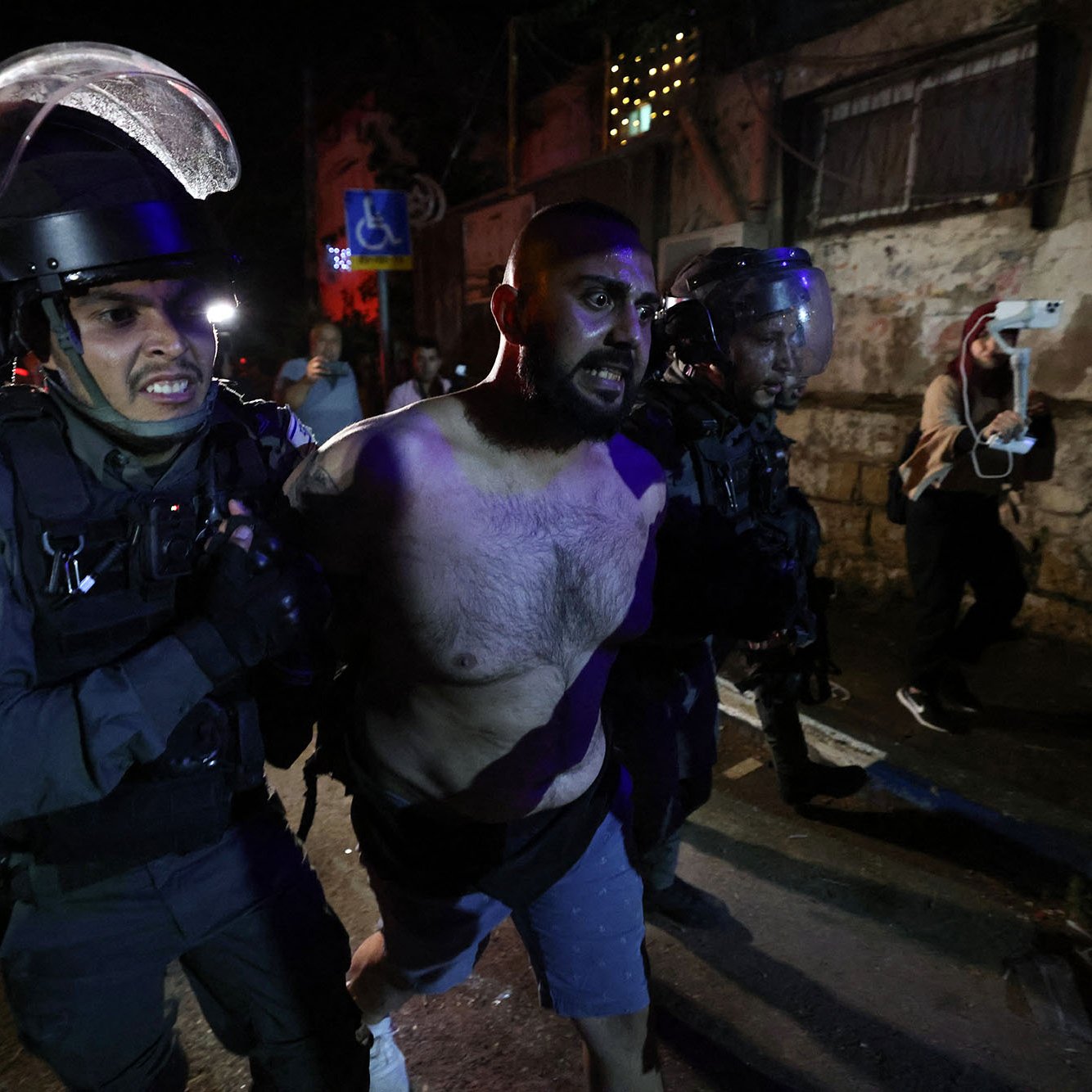 تظهر الصورة مجموعة من رجال الشرطة الذين يقومون باعتقال رجل. يبدو الرجل عاري الصدر ويظهر عليه علامات التوتر أو المقاومة. تحيط به عناصر من الشرطة يرتدون معدات حماية، ويبدو أنهم يتخذون إجراءات للسيطرة عليه. الخلفية توحي بأن المكان ربما كان شاهداً على مظاهر من التوتر أو الاضطراب، مع وجود إضاءة خافتة تشكل جوًا متوترًا.