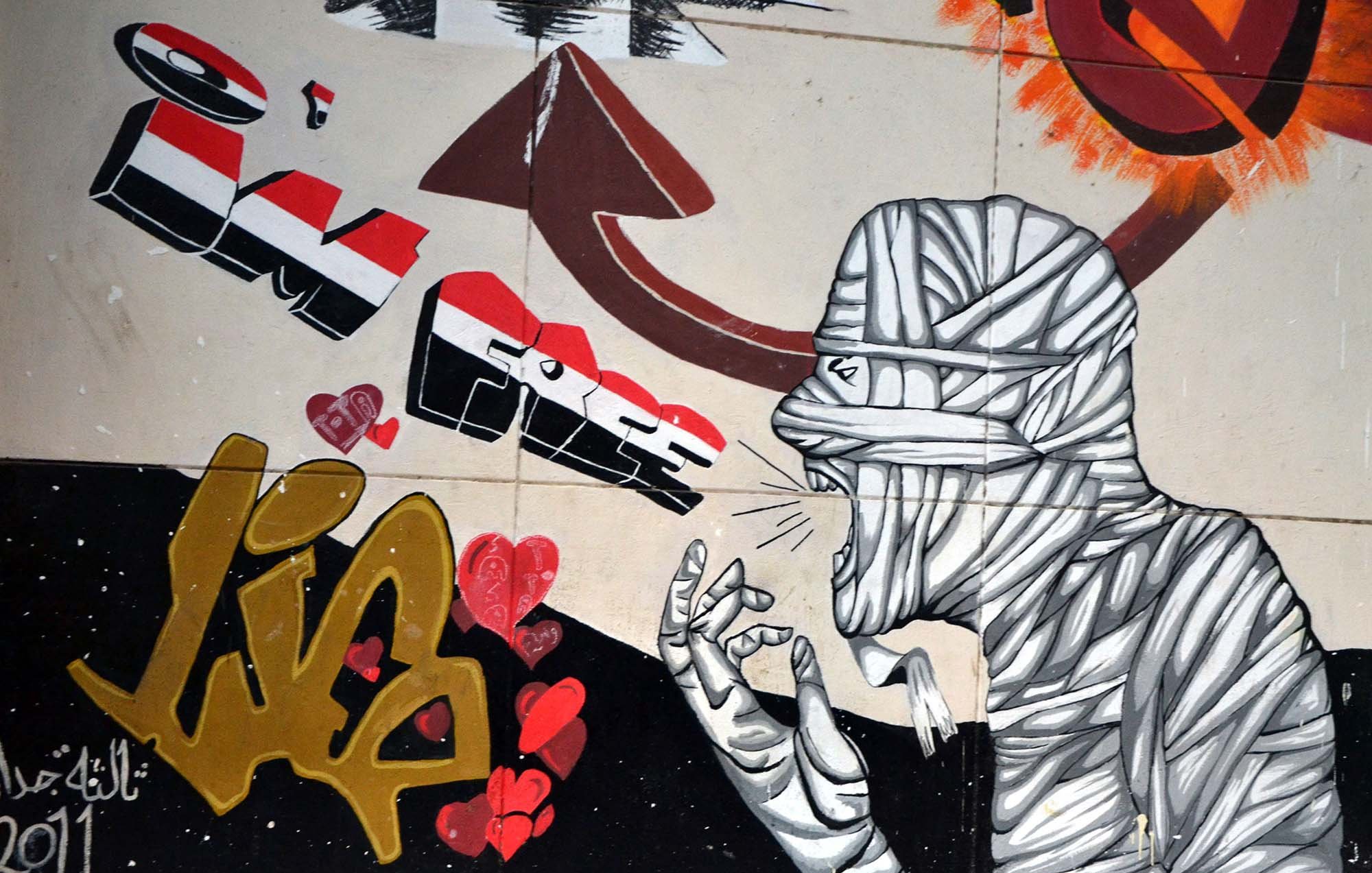 L'image présente une œuvre de street art qui illustre une figure humaine entourée de graffitis colorés. La silhouette est représentée de manière stylisée, avec un design en noir et blanc, et semble crier ou exprimer une forte émotion. Des cœurs rouges sont dispersés autour, ajoutant une touche de passion. À côté, le mot "Lib" est écrit en lettres gold avec une certaine flamboyance, peut-être symbolisant la liberté. L'ensemble évoque un message fort, reliant des thèmes d'expression et d'émotion à un contexte historique ou social.