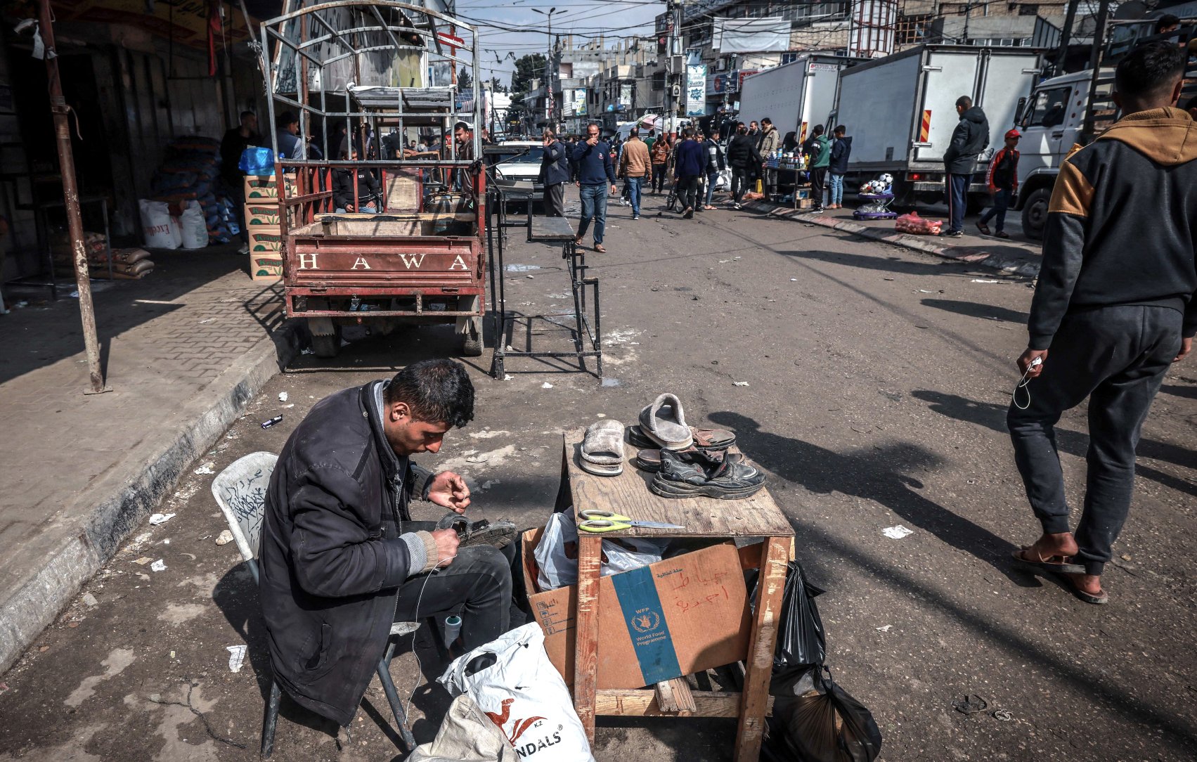 L'image montre une scène de rue animée. Au premier plan, un homme est assis sur une chaise, concentré sur sa tâche. Il répare des chaussures, utilisant des outils comme des ciseaux et des filaments. Devant lui, une table supporte des chaussures à réparer et un sac. À l'arrière-plan, on peut voir des personnes marchant le long d'une rue entourée de camions et de petites échoppes. L'atmosphère semble vivante, avec des éléments de la vie quotidienne qui se déroulent autour de lui.
