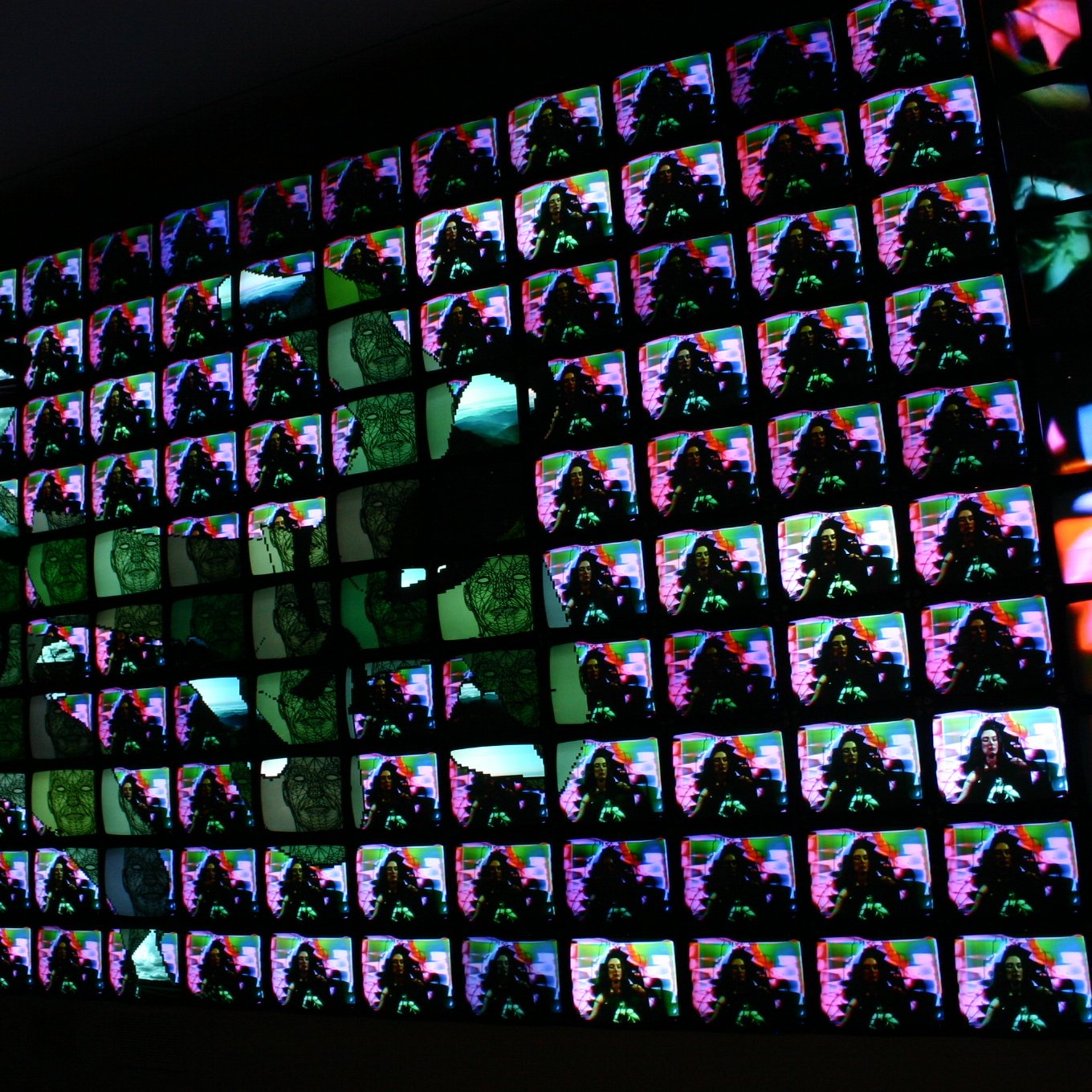 تصویر شامل یک دیوار بزرگ است که از تعداد زیادی تلویزیون کوچک تشکیل شده است. هر یک از این تلویزیون‌ها تصاویری با رنگ‌های متنوع و الگوهای پیچیده را نمایش می‌دهند. به نظر می‌رسد که تصاویری از انسان‌ها یا اشیاء در پس‌زمینه وجود دارد که در کنار هم نمایی جذاب و هنری ایجاد کرده‌اند. ترکیب این تصاویر باعث ایجاد یک تجربه بصری زنده و دینامیک می‌شود.