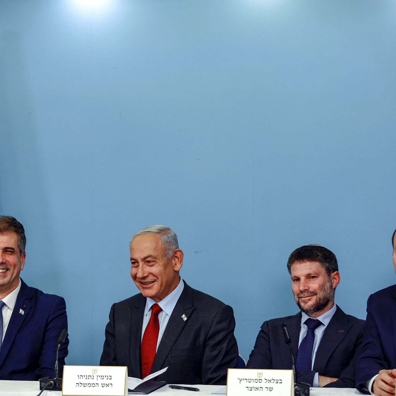 La imagen muestra a un grupo de cuatro personas sentadas alrededor de una mesa en un entorno oficial. Al fondo, se puede ver una bandera de Israel. Las personas parecen estar en un evento o una conferencia, y todos tienen expresiones serias pero amigables. Están vestidos de manera formal. La escena sugiere un contexto político o gubernamental.