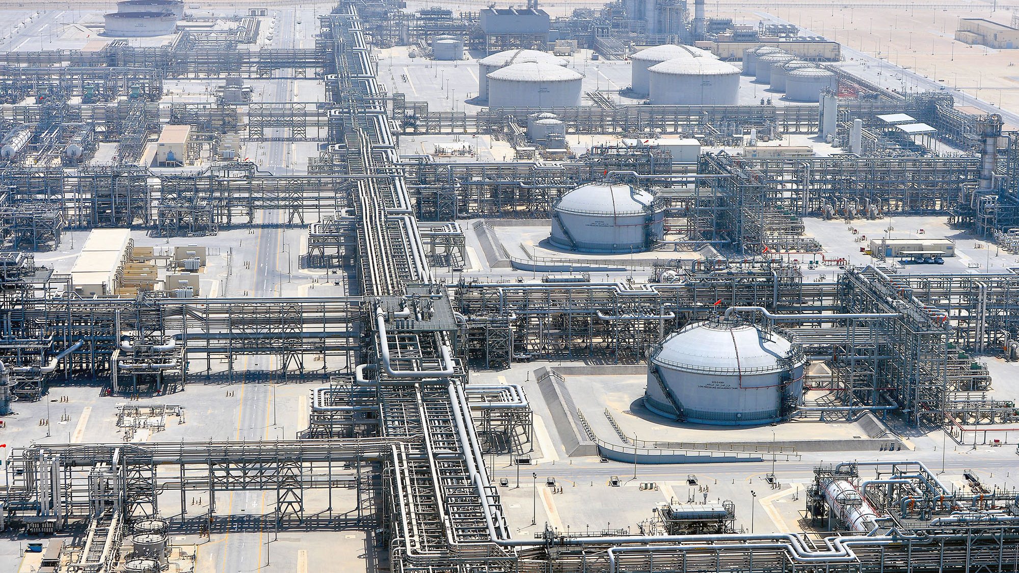 L'immagine mostra un impianto industriale, probabilmente una raffineria o un complesso di estrazione di prodotti petroliferi. Si possono vedere grandi serbatoi rotondi e una vasta rete di tubazioni che si snodano attraverso il sito. L'area circostante appare deserta, con una superficie di sabbia o terra arida. Sullo sfondo si intravedono strutture alte, che potrebbero essere torri di raffineria o impianti di trattamento. L'intero complesso è ben organizzato, con una disposizione sistematica delle tubazioni e dei serbatoi.