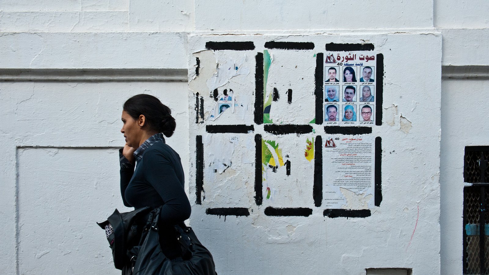 L'image montre une femme marchant le long d'un mur blanc. Elle porte un haut noir et une écharpe. Le mur derrière elle est décoré de divers papiers affichés, y compris des portraits de plusieurs personnes et des textes en arabe. L'atmosphère de la scène semble urbaine et vivante, avec un contraste entre la femme et le mur.