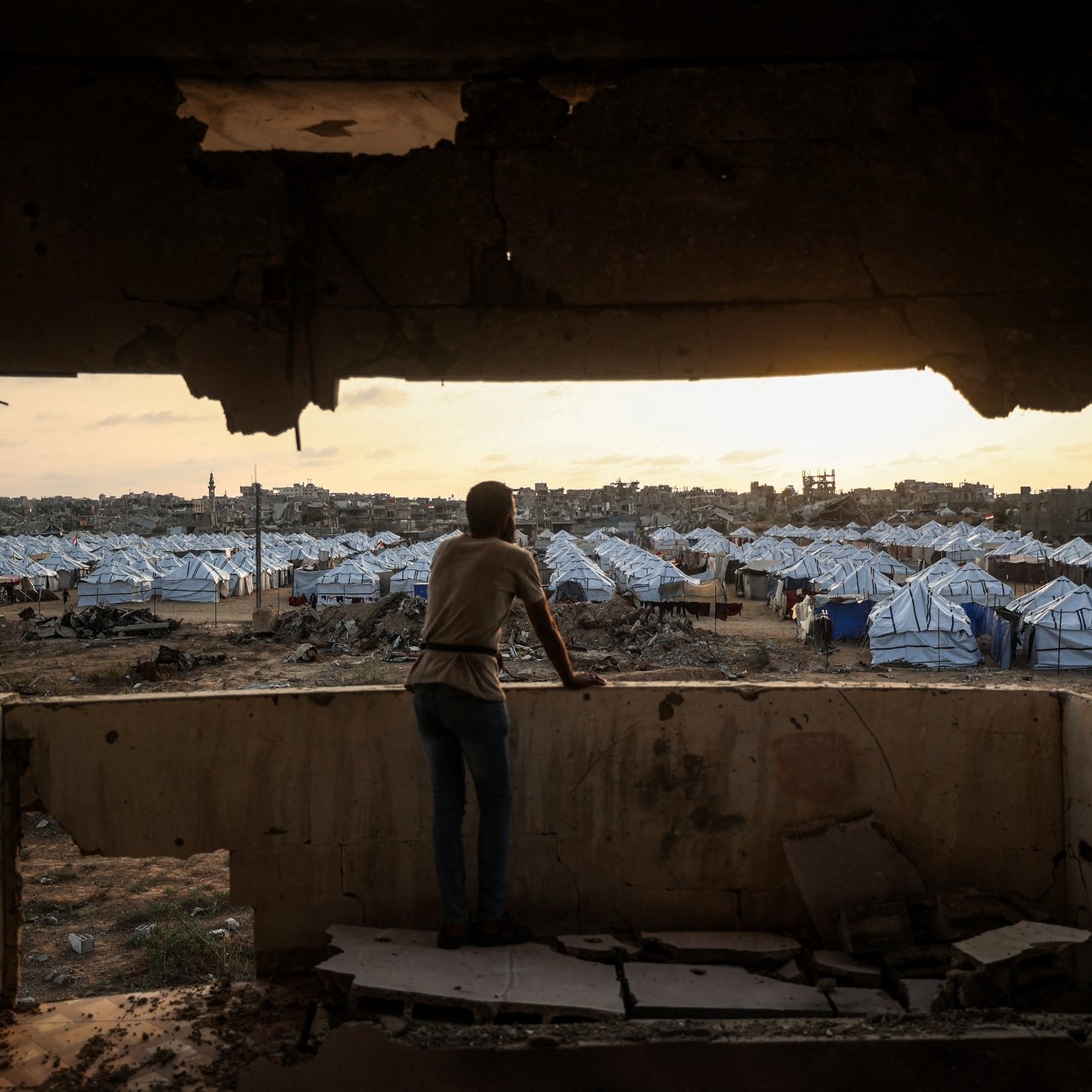 Un jeune observe un camp de tentes depuis une fenêtre endommagée au crépuscule.