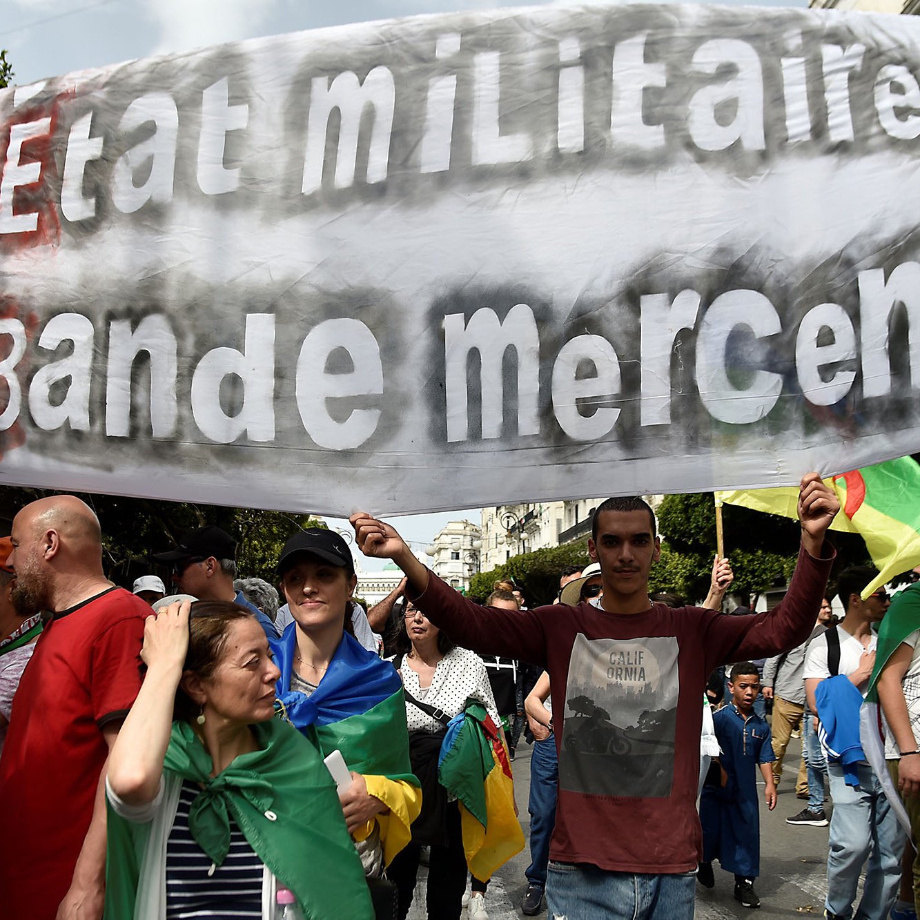 L'image montre une manifestation où des gens tiennent une grande banderole. Sur celle-ci, on peut lire "Ni État militaire, ni Bande mercenaire", ce qui reflète un message de revendication politique. Les manifestants semblent porter des drapeaux et exprimer des sentiments de dissentement. L'ambiance est mobilisée, avec des personnes de différents âges rassemblées pour une cause commune.
