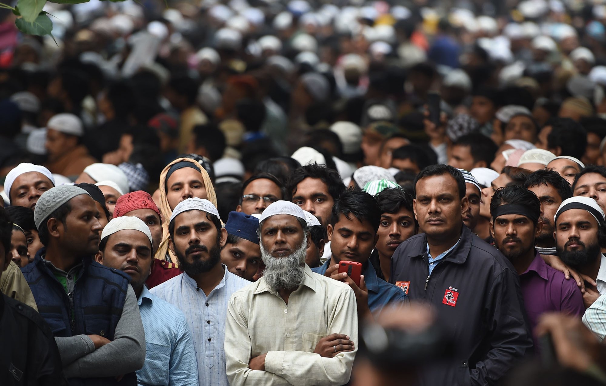 The image shows a large crowd of people, primarily men, gathered closely together. Many of the individuals are wearing traditional Islamic attire, including caps (taqiyah or kufi). The expressions on their faces suggest a mix of focus and anticipation. The background is filled with more attendees, creating a sense of a significant event or gathering. The atmosphere appears to be one of unity and collective participation.
