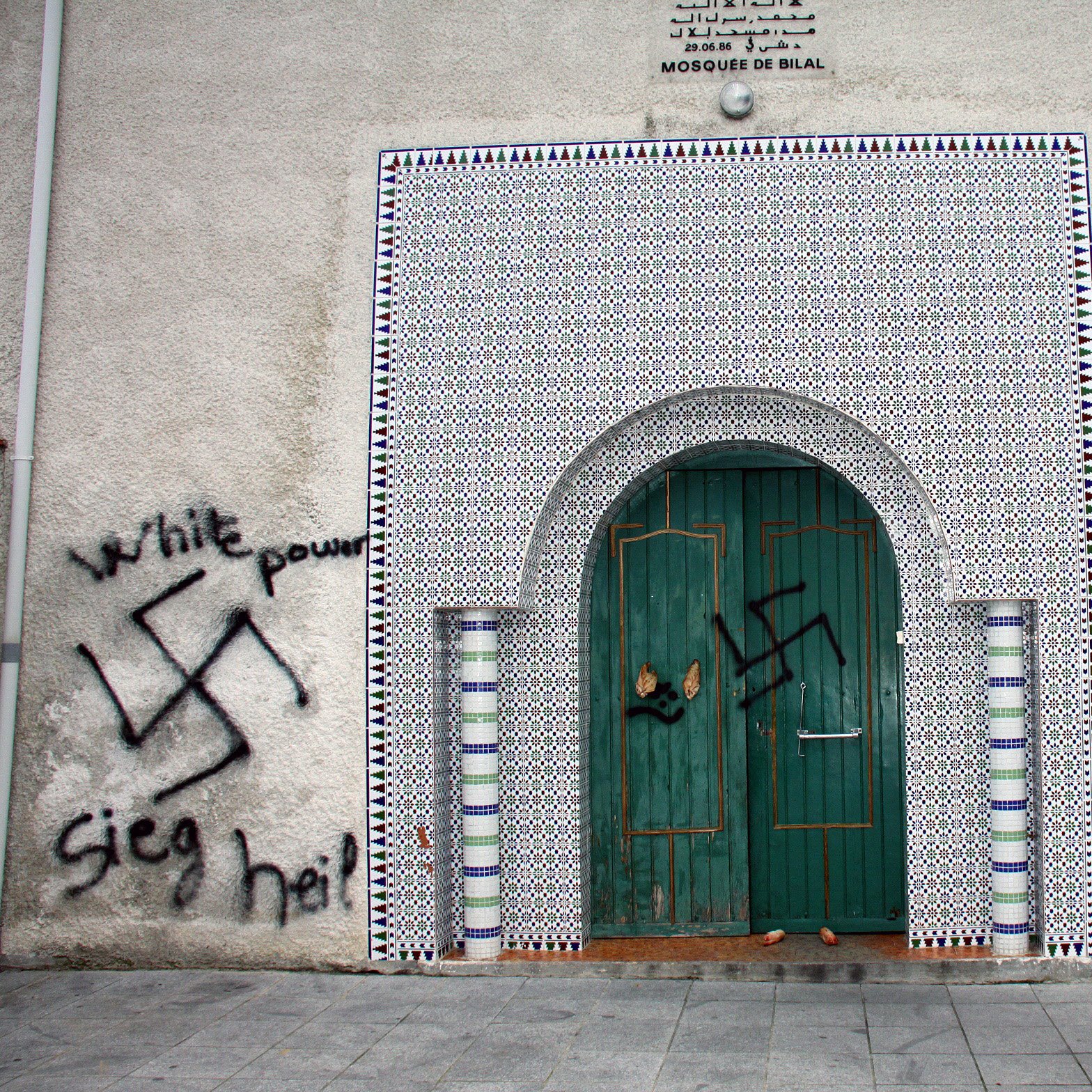 این تصویر نمایی از دیواری را نشان می‌دهد که در آن نمادهای نژادپرستی و خشونت، از جمله علامت هیکل و نوشته‌هایی به زبان‌های مختلف، Spray شده است. در بالای درب ورودی، طراحی‌های اسلامی به چشم می‌خورد که نشان‌دهنده یک مسجد است. دیوار به طور کلی نشان‌دهنده تضاد بین هنر و پیام‌های نفرت است.