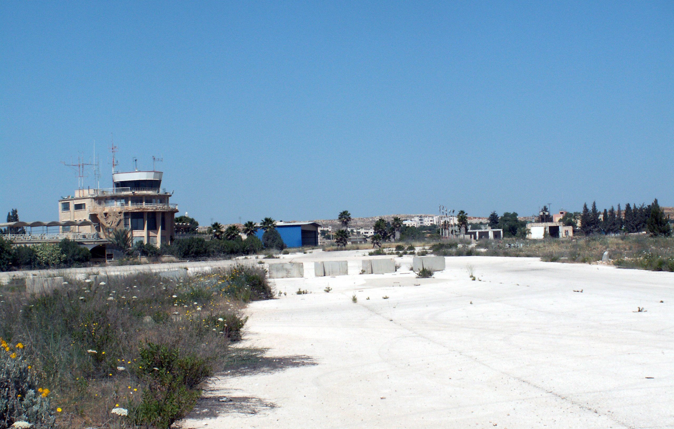 La imagen muestra un paisaje de un área desierta, posiblemente un antiguo aeropuerto o base aérea. En el fondo se observa una torre de control o edificio administrativo, rodeado de vegetación y estructuras en desuso. El suelo es de color claro y parece estar cubierto de hierbas y flores silvestres, lo que sugiere abandono. El cielo es de un azul despejado, lo que le da un contraste al paisaje.