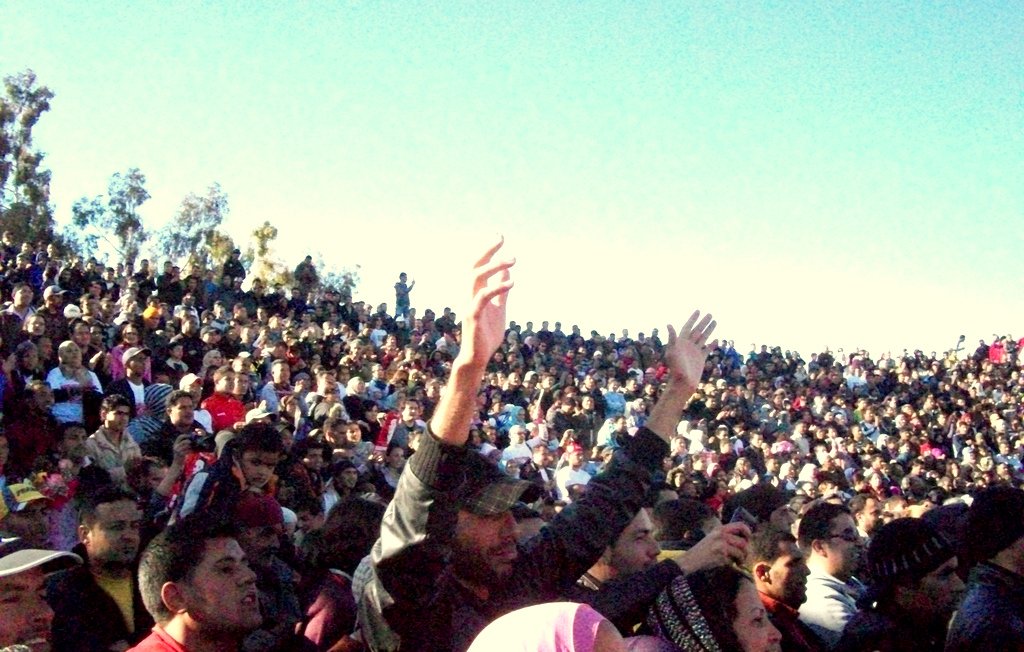 L'image montre une vaste foule de personnes rassemblées dans un espace ouvert. On peut voir des milliers de spectateurs, certains levant les bras en l'air, clairement engagés et enthousiastes. L'arrière-plan est constitué de buissons et d'arbres, tandis que le ciel est bleu et dégagé. L'atmosphère semble festive et dynamique, suggérant un événement important ou un concert.
