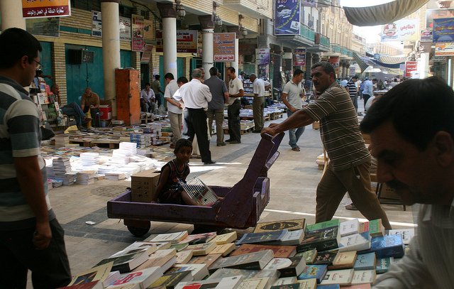 L'image montre un marché animé, probablement un souk, avec des étals de livres disposés au sol. On peut voir plusieurs personnes se déplacer dans la rue, certains feuilletant des livres. Un homme pousse un chariot, où un enfant est assis, observant autour de lui. Les bâtiments en arrière-plan suggèrent une architecture locale, et l'ambiance paraît chaleureuse et vivante.