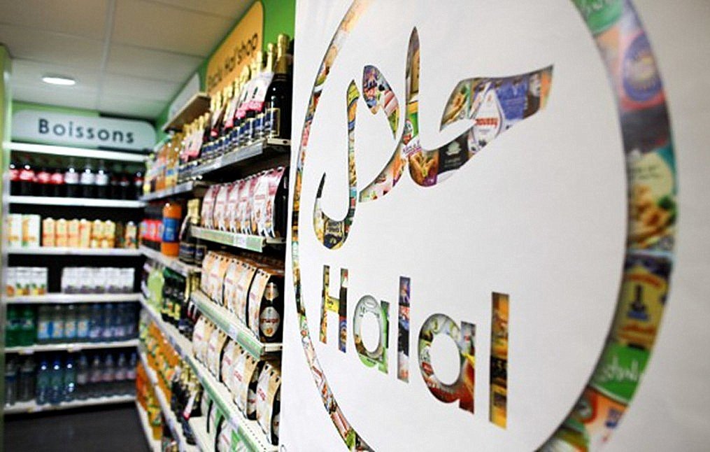 L'image montre une vue intérieure d'un magasin avec une étagère remplie de divers produits alimentaires et boissons. Au premier plan, il y a un grand logo représentant le mot "Halal" en lettres stylisées. Ce logo est intégré dans un design créatif qui semble inclure des images de produits alimentaires. L'éclairage est clair, et l'ambiance générale de la scène est celle d'un espace de vente organisé et vibrant.