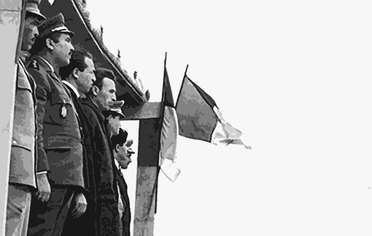 L'image montre un groupe de militaires et de dignitaires, probablement lors d'une cérémonie officielle. Ils se tiennent sur une estrade, certains en uniforme avec des médailles, tandis que d'autres portent des vêtements civils. En arrière-plan, on peut voir des drapeaux, dont un drapeau français, indiquant un contexte lié à la France ou un événement officiel. Les soldats se tiennent en position de parade, face à l'estrade. L'ambiance semble solennelle.