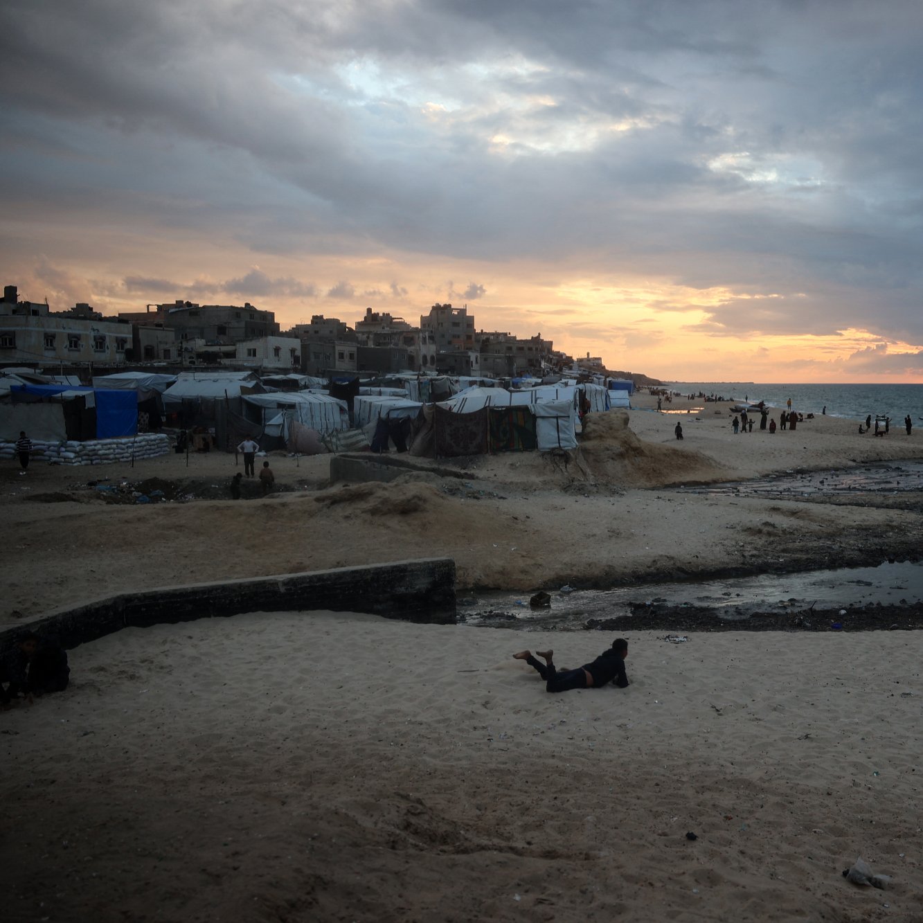 تصویر نشان‌دهنده‌ی یک ساحل است که در آن آسمان پهناوری با ابرهای تیره و روشن به تصویر کشیده شده است. خورشید در حال غروب است و نور ملایمی بر روی آب دریا و شن‌های ساحل می‌تابد. در سمت چپ تصویر، برخی چادرها و سازه‌هایی دیده می‌شود که نشان‌دهنده‌ی سکونتگاه‌های موقت هستند. همچنین، در دوردست، افرادی در حال حرکت و فعالیت هستند. این تصویر حال و هوای خاصی از مکان‌های ساحلی همراه با چالش‌های اجتماعی و محیطی را منتقل می‌کند.