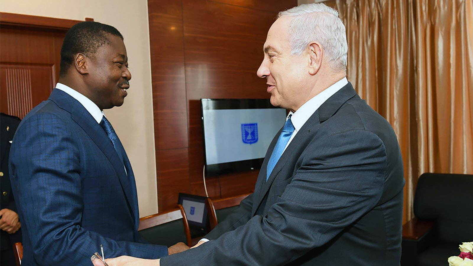 الصورة تظهر رجلين يتصافحان في مناسبة رسمية. يبدو أن أحدهما يرتدي بدلة زرقاء داكنة ويبتسم، بينما الآخر يرتدي بدلة سوداء مع ربطة عنق زرقاء. الخلفية تحتوي على ستائر مصنوعة من القماش الفاخر، ويتواجد هناك جهاز كمبيوتر محمول على الطاولة خلفهم. الجو في الصورة يوحي بالود والترحيب.