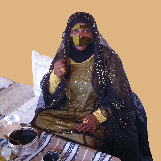 این تصویر یک زن را نشان می‌دهد که در لباس و پوشش سنتی خود نشسته است. او چهره‌اش را با نوار مشکی پوشانده و چشم‌هایش نمایان است. لباس او شامل رنگ‌های تیره و زینت‌های طلایی است که درخشندگی خاصی دارد. زن در حال نشستن بر روی یک سجاده یا پارچه با خطوط افقی است و در کنار او چند وسیله مانند ظرف‌های کوچک دیده می‌شود. زمینه تصویر نیز به رنگ قهوه‌ای ملایم است و به عکس حس گرم و جذابی می‌دهد.