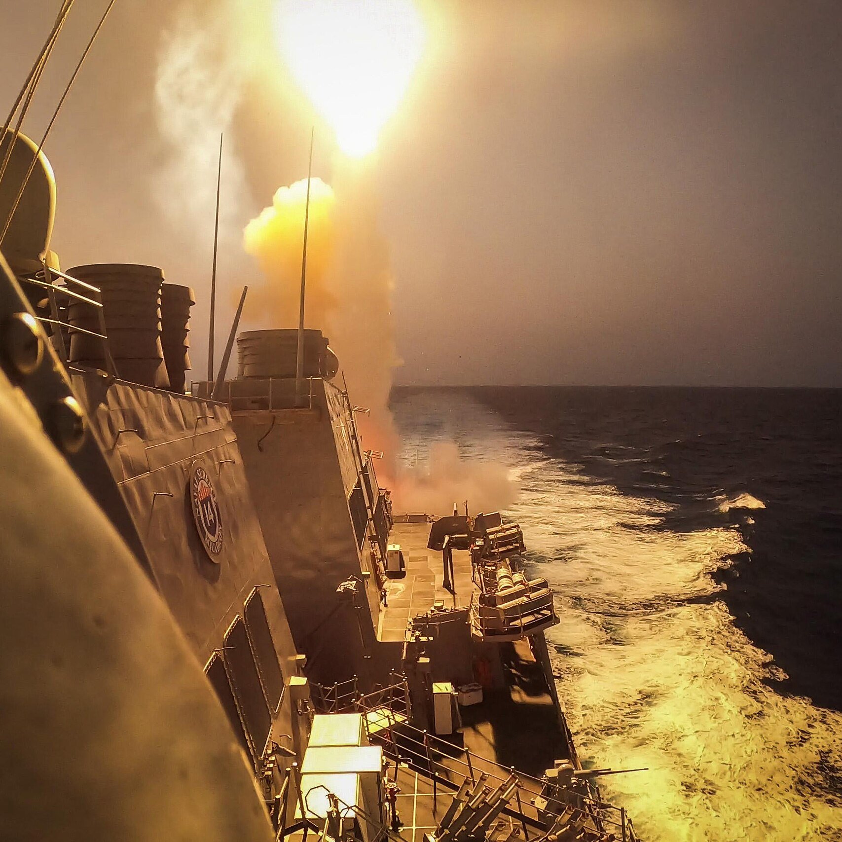 L'immagine mostra una scena marittima in cui un missile viene lanciato da una nave da guerra. Si vede il missile che viene sparato con un forte bagliore e un'esplosione di fumi dietro di esso. In primo piano si possono notare le strutture della nave e il mare in movimento, evidenziato da schiuma bianca. Il cielo appare velato e si intuisce un momento di grande intensità e potenza militare.