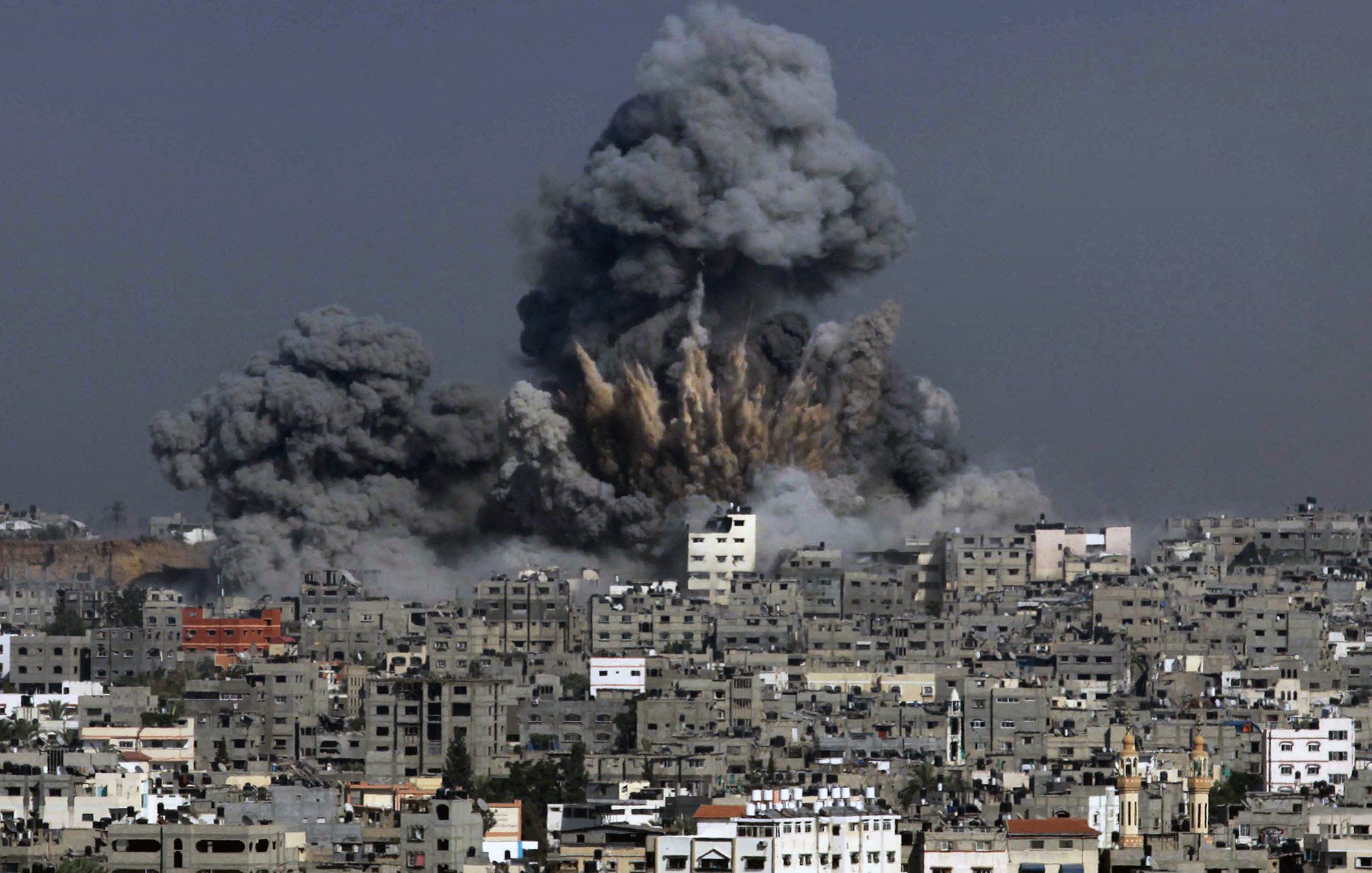 الصورة تظهر مشهدًا لمدينة تعرضت لتفجير، حيث تندلع سحب كثيفة من الدخان الأسود في السماء. يظهر تأثير الانفجار في المباني المحيطة، حيث تبرز آثار الدمار، بينما يبدو أن السماء ملبدة بالغيوم الناتجة عن الانفجار. المدينة تبدو كثيفة البنايات مع تشوهات واضحة نتيجة الأحداث المدمرة.