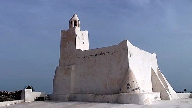 L'image montre un bâtiment de couleur blanche, typiquement de style architectural méditerranéen. Il semble être une petite structure, possédant une forme irrégulière avec des murs épais et angulaires. À son sommet, il y a une cloche ou un petit clocher. L'arrière-plan est plutôt clair, avec un ciel bleu ou nuageux qui met en valeur la luminosité de l'édifice. Ce type de construction pourrait évoquer des lieux historiques ou religieux, typiques de certaines régions côtières.
