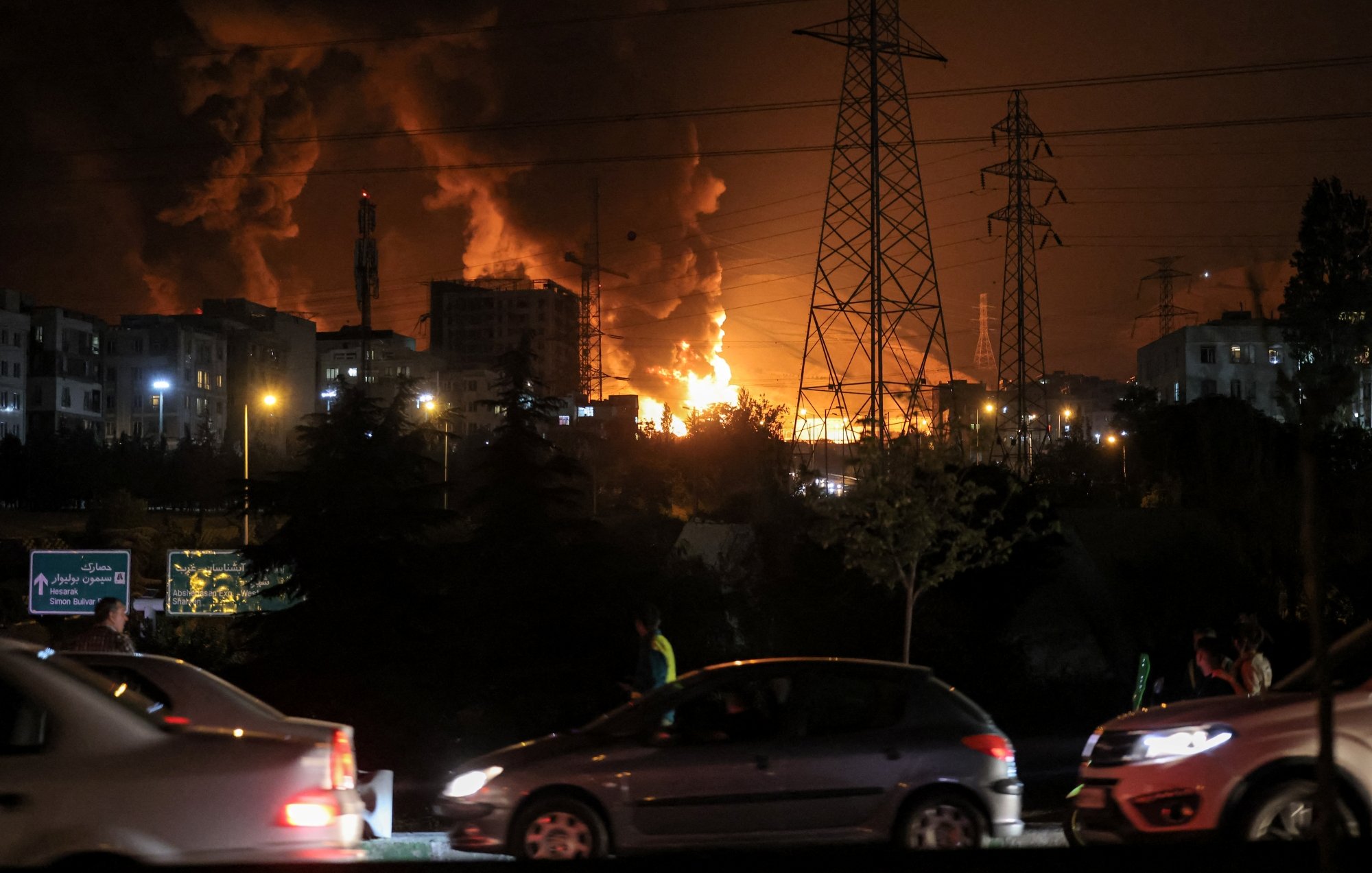 Flammes illuminent la nuit, voitures passent près d'une scène de chaos urbain.