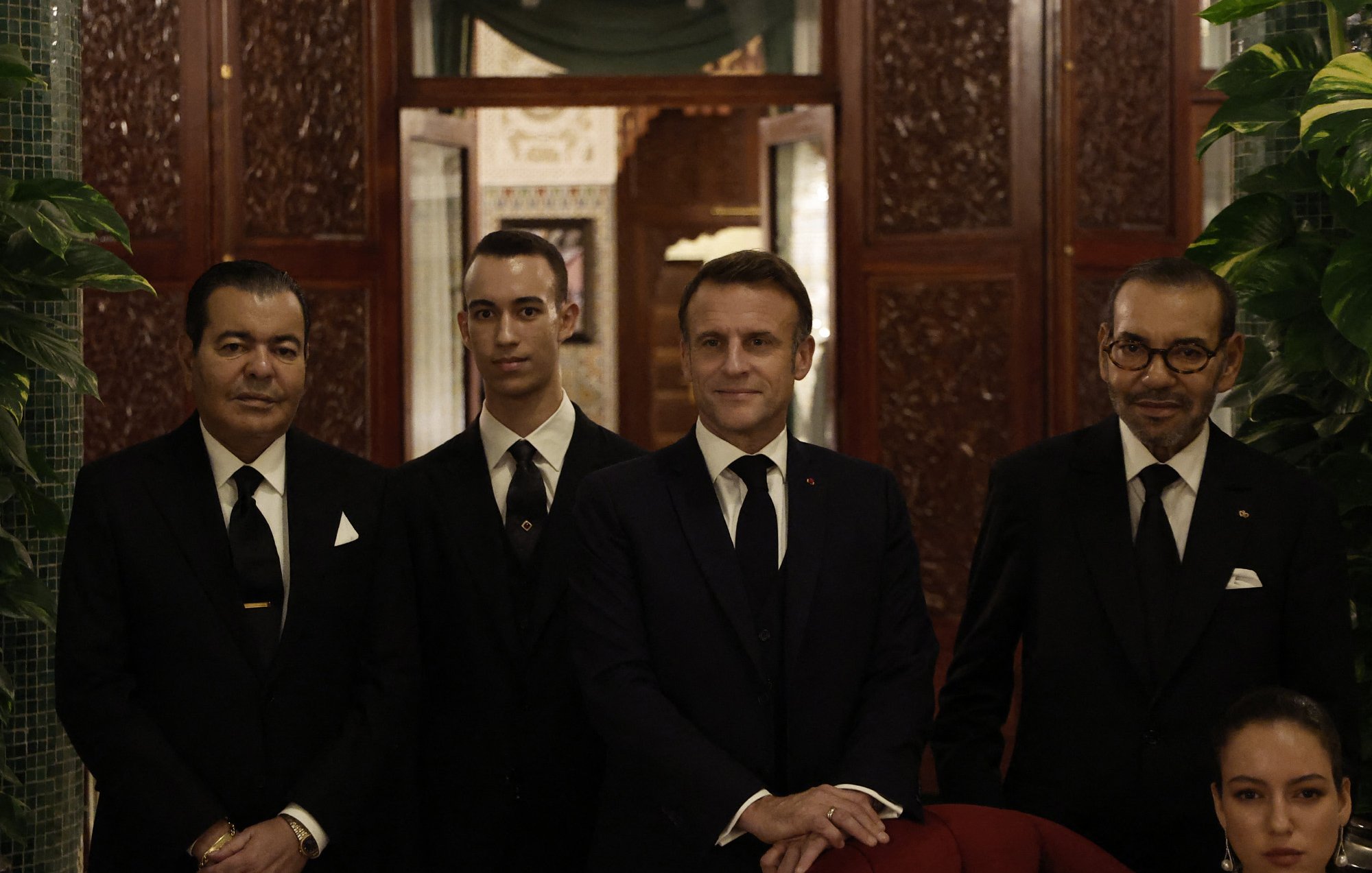 يظهر في الصورة مجموعة من الرجال يرتدون بدلات رسمية سوداء. يقع شخص في الوسط يبدو أنه في موقع قيادي أو مهم، حيث يرتدي بدلة داكنة وربطة عنق. إلى جانبه، هناك شخص آخر يرتدي أيضًا بدلة، بالإضافة إلى رجلين آخرين على الجانبين. الخلفية تتضمن تفاصيل معمارية جميلة، مع نقوش وزخارف تقليدية. هناك نباتات خضراء تضيف لمسة طبيعية إلى المشهد. 분위기 يظهر احتفاليًا ورسميًا.