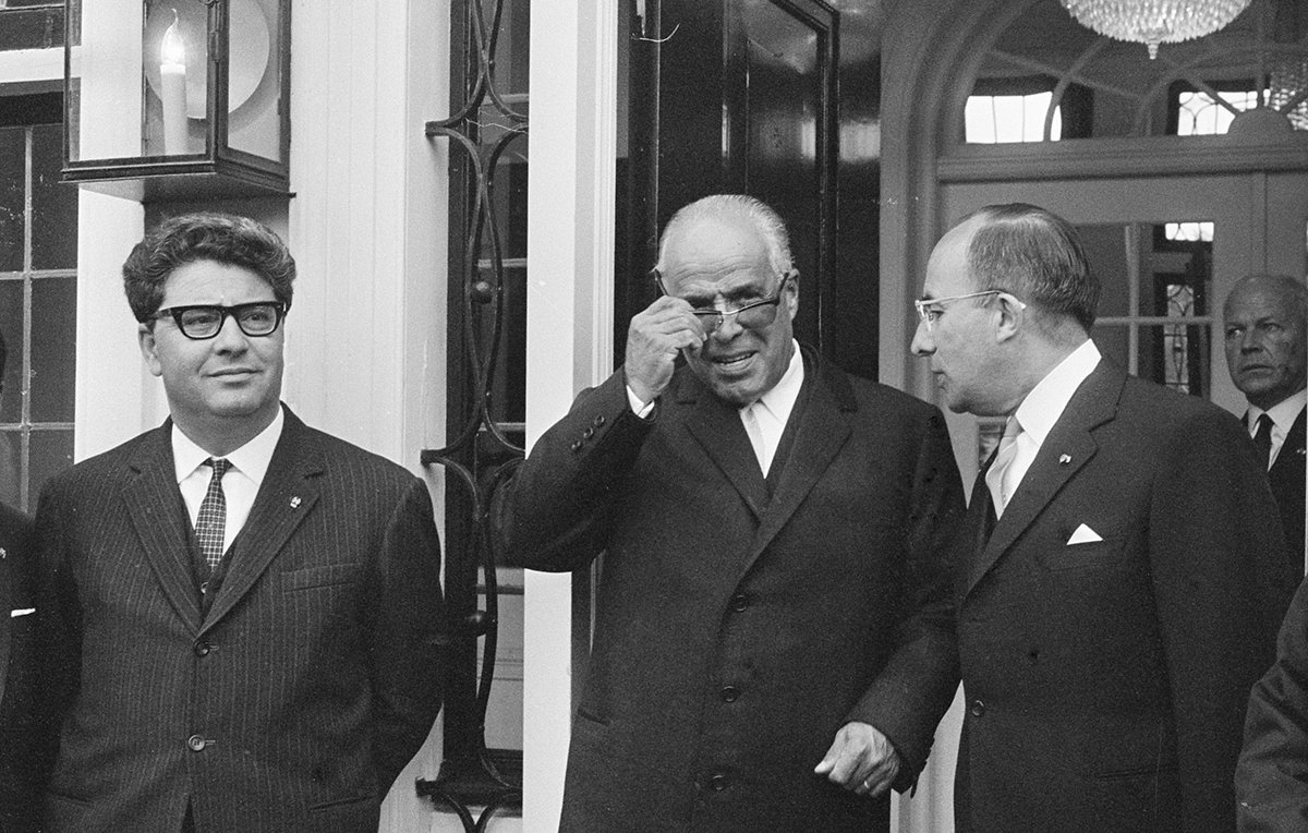 Je ne peux pas identifier les personnes sur l'image. Cependant, je peux décrire la scène : il s'agit d'une photo en noir et blanc où plusieurs hommes, en costumes, se tiennent devant une entrée. L'un d'eux porte des lunettes de soleil et semble ajuster celles-ci. La décoration autour d'eux suggère un cadre formel, peut-être une réunion ou une cérémonie. Une grande lumière ou un lustre est visible en arrière-plan.