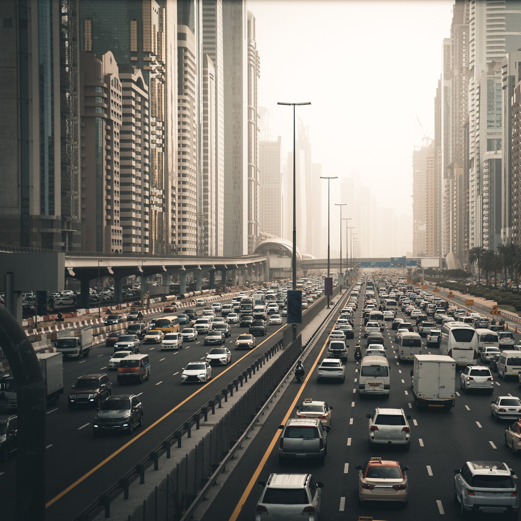 این تصویر نمایی از یک خیابان شلوغ در یک شهر مدرن را نشان می‌دهد. در سمت چپ، ساختمان‌های بلند و مرتفع با معماری‌های معاصر دیده می‌شوند. خیابان مملو از خودروهاست که در دو طرف حرکت می‌کنند. ترافیک به وضوح قابل مشاهده است و برخی خودروها در حالت توقف به نظر می‌رسند. در زمینه، مه یا گرد و غباری به چشم می‌خورد که باعث کاهش وضوح چشم‌انداز می‌شود. فضای کلی تصویر حس شلوغی و پویا بودن شهر را منتقل می‌کند.