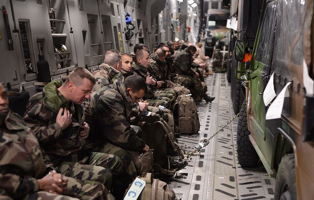 L'image montre un groupe de soldats assis à l'intérieur d'un véhicule militaire ou d'un avion de transport. Ils portent des uniformes camouflage et sont équipés de sacs à dos. Certains semblent attentifs et concentrés, tandis que d'autres sont plus détendus. Le cadre est sombre et fonctionnel, typique des espaces militaires, avec des sièges alignés sur les côtés et un véhicule militaire garé à l'intérieur.