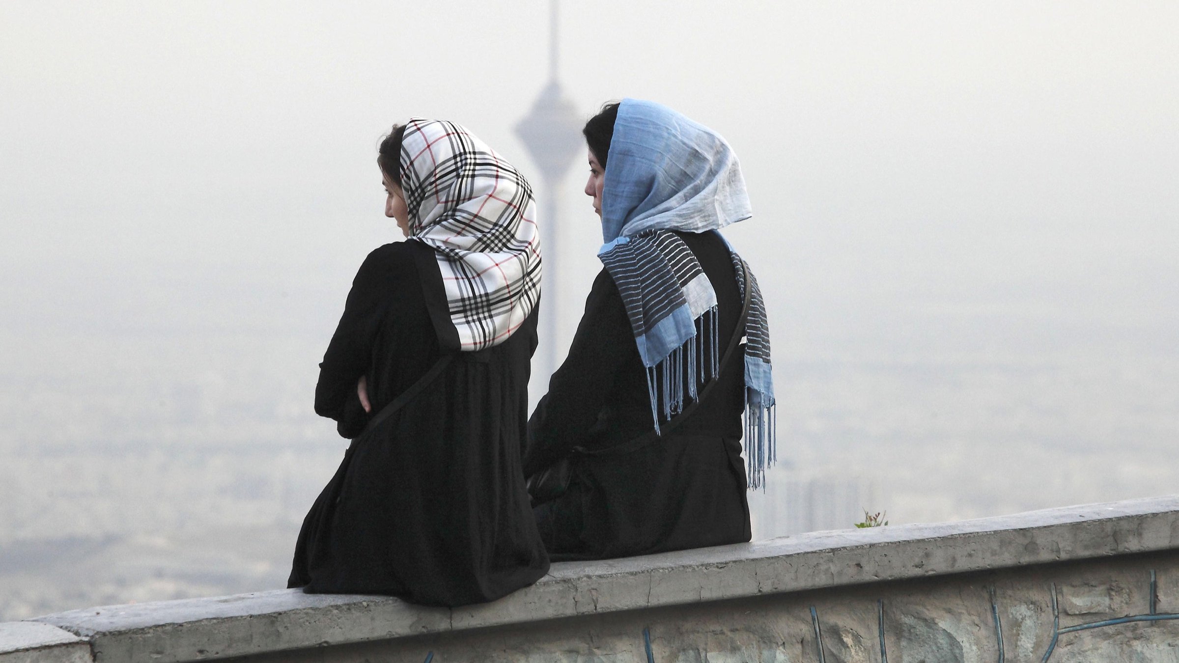 L'image montre deux femmes assises sur un mur, avec le dos tourné à la caméra. Elles portent des vêtements longs et des foulards, l'un d'eux étant à motifs. En arrière-plan, on aperçoit une ville embrumée avec ce qui semble être une tour, ajoutant un aspect urbain à la scène. L'atmosphère est paisible, avec une lumière douce, suggérant un moment de réflexion ou de contemplation.