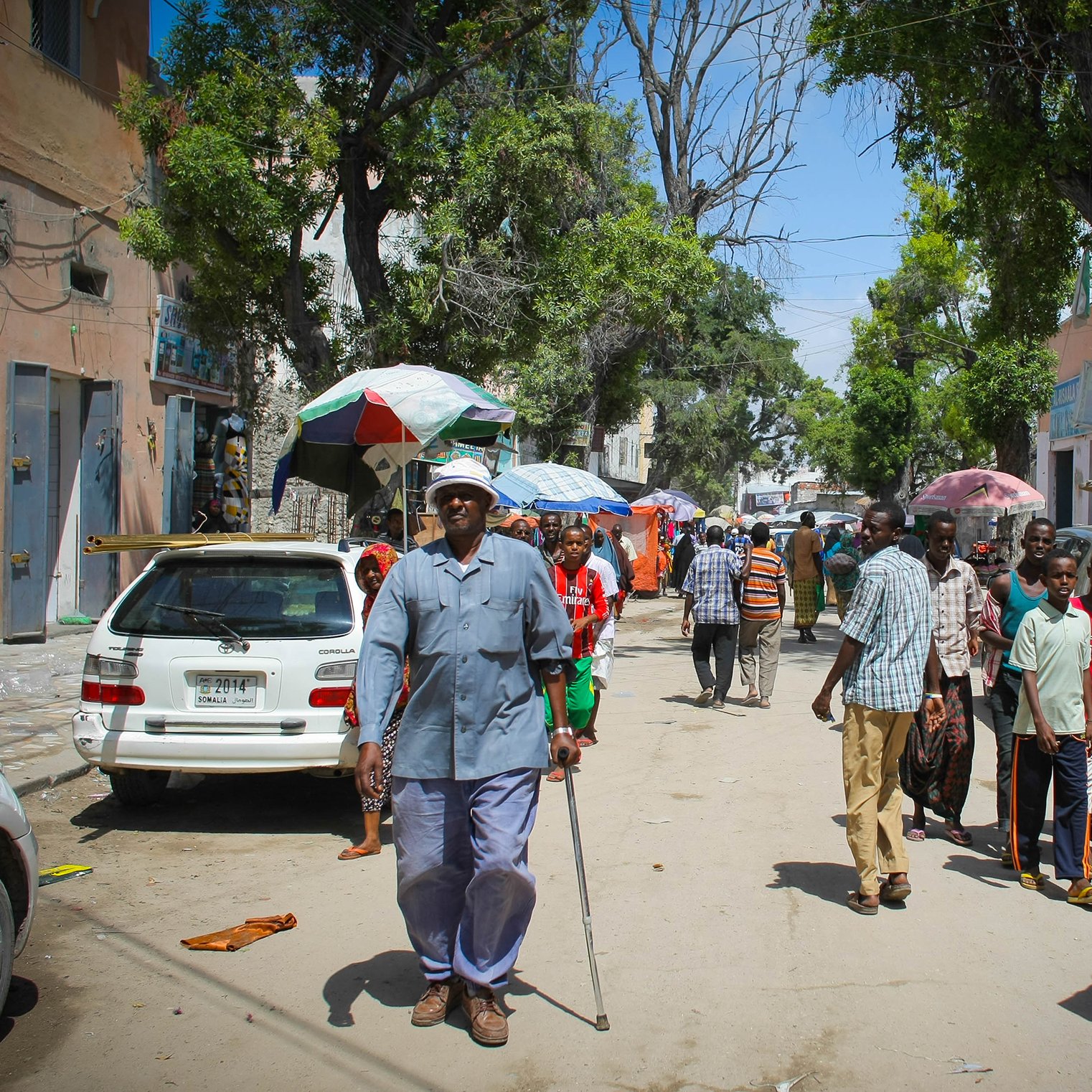 تظهر الصورة شارعا في منطقة حيوية، حيث يسير مجموعة من الأشخاص في اتجاهات مختلفة. في المقدمة، يوجد رجل مسن يستخدم عكازا للمشي، بينما يتواجد أشخاص آخرون في الخلفية، بعضهم يتحدث والبعض الآخر يسير. هناك سيارات متوقفة على الجانبين، وتظلل الأشجار والشمس المكان. يبدو أن الجو مشمس والناس يقومون بأعمالهم اليومية.