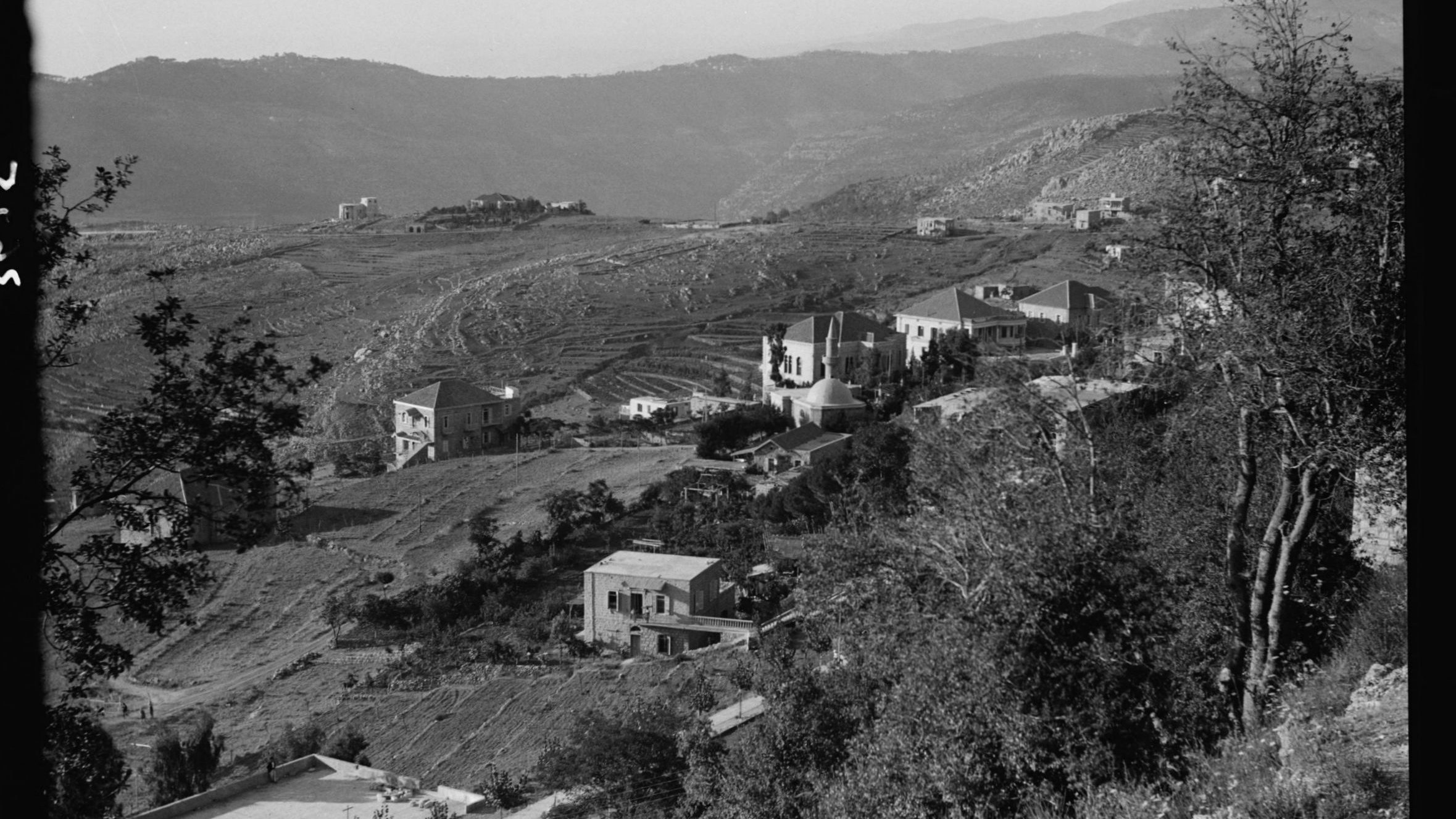 L'image montre un paysage montagneux avec des collines qui s'étendent à l'horizon. On peut apercevoir plusieurs maisons éparpillées sur les flancs des collines, entourées de végétation. La scène semble être prise en noir et blanc, ce qui lui donne un aspect nostalgique. Les terrains agricoles sont visibles, indiquant une zone rurale. Les montagnes en arrière-plan ajoutent une profondeur à la composition paysagère.