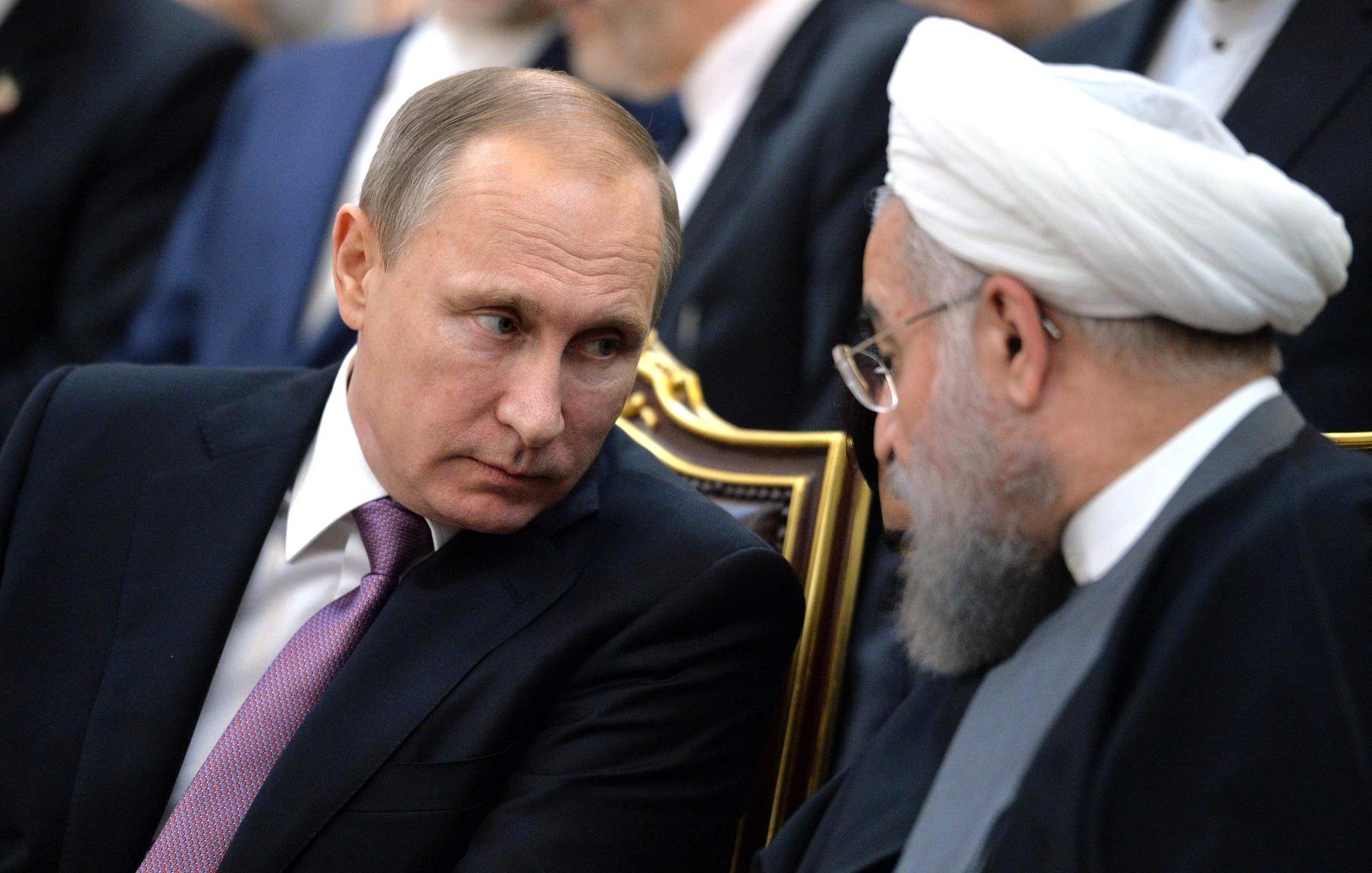 The image depicts two men engaged in a conversation. One is seated with a serious expression, wearing a dark suit and a light-colored tie. The other man, also seated, has a beard and wears traditional clerical attire, including a white turban. They appear to be in a formal setting, possibly during a meeting or event, with other individuals visible in the background. The focus is on their interaction and expressions.