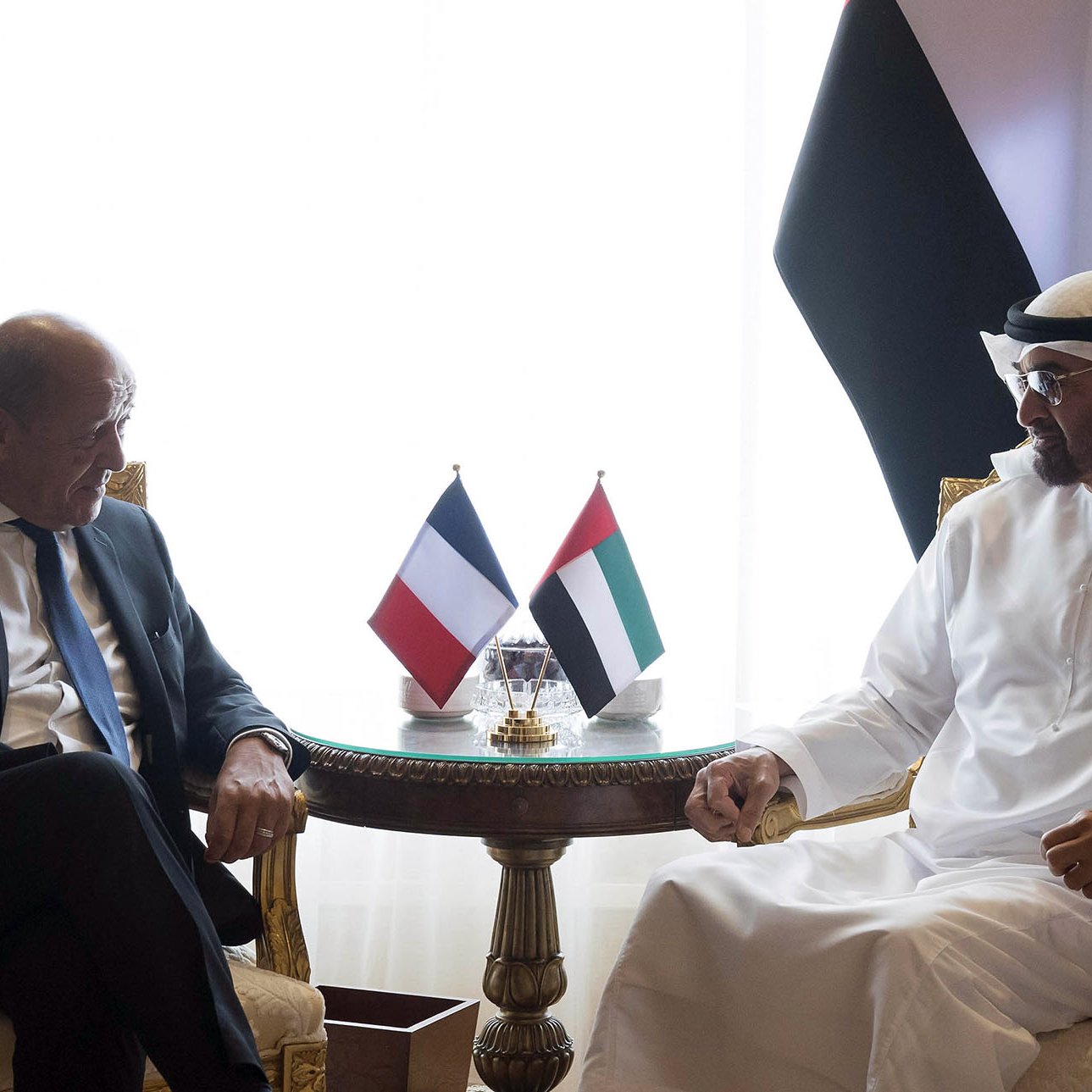 تظهر الصورة لقاءً بين شخصين يجلسان في غرفة تتسم بالأناقة. الشخصان يجلسان على كراسي فاخرة أمام طاولة صغيرة، حيث توجد علمان. أحد العلمين يعود إلى فرنسا والآخر يعود إلى الإمارات العربية المتحدة. يبدوان الشخصان في حالة حوار ودية، مع تعابير تشير إلى الانفتاح والنقاش. الإضاءة في الغرفة دافئة، والستائر خلفهم تضيف لمسة من الفخامة.