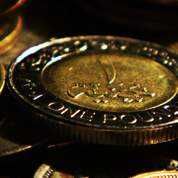 L'image montre plusieurs pièces de monnaie, avec une pièce de un pound en avant-plan. La pièce est en métal, et on peut voir des inscriptions en arabe sur sa surface, ainsi que les mots "ONE POUND". L'arrière-plan est flou, composé de plusieurs autres pièces de monnaie qui ajoutent à la richesse visuelle de la composition. Les pièces semblent avoir un éclat doré, ce qui attire l'attention sur le sujet principal.