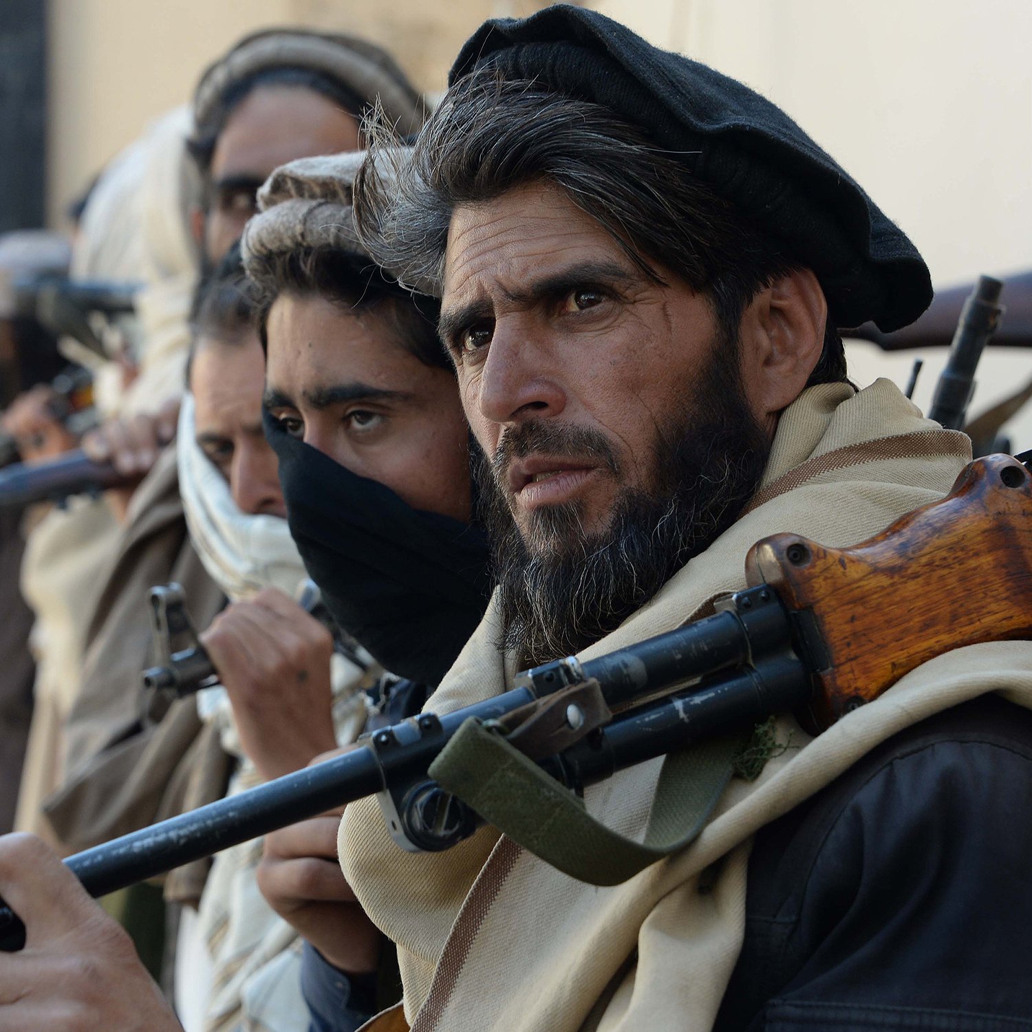 The image features a group of individuals standing closely together, each holding firearms. They appear to be dressed in traditional clothing, with some wearing headscarves and others with their faces partially obscured. The focus is on one man in the foreground, who has a serious expression and a well-groomed beard. The background is unfocused, but it suggests a rugged, possibly desert-like environment. The overall mood conveys tension or readiness.