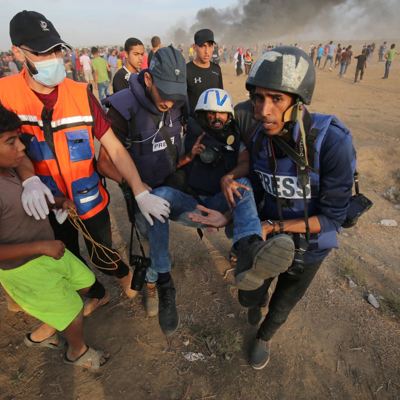 L'image montre une scène dramatique où plusieurs personnes, dont des journalistes, interviennent dans une situation d'urgence. Un homme blessé est transporté par des secouristes, tandis qu'un jeune garçon semble les observer. En arrière-plan, il y a des fumées, probablement d'incendies, et une foule rassemblée. L'environnement semble chargé d'émotion et de tension, évoquant des événements majeurs ou des conflits.