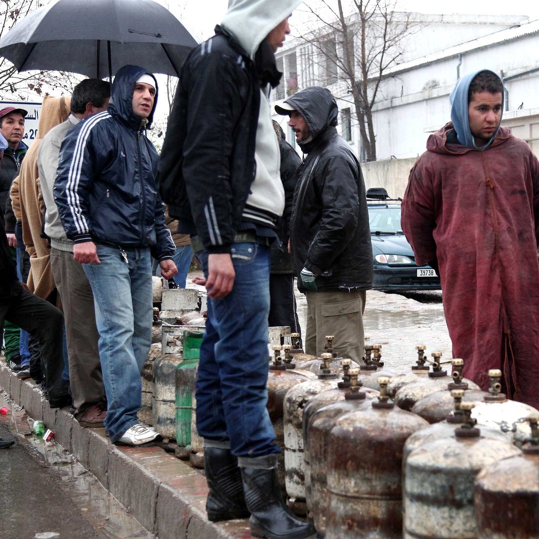 L'image montre un groupe de personnes rassemblées sous la pluie, attendant près de bouteilles de gaz. Certains portent des vêtements imperméables, comme des vestes et des manteaux, tandis que d'autres se protègent de la pluie avec des parapluies. L'atmosphère semble sombre et humide, suggérant une situation de manque ou d'attente. En arrière-plan, des voitures sont visibles, ainsi que des bâtiments. Les gens semblent engagés dans une activité collective, probablement en attendant d'acheter du gaz.