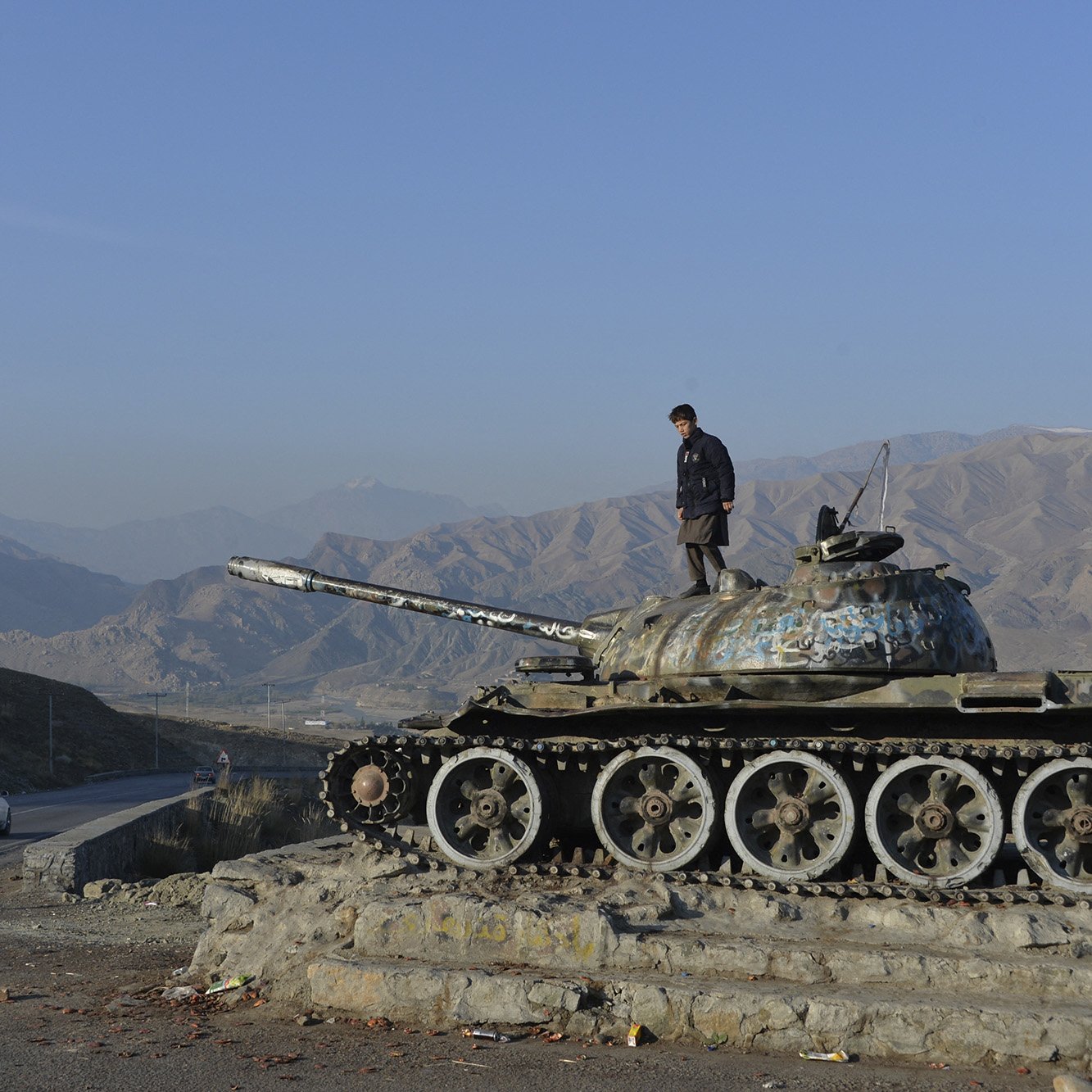 L'immagine mostra un carro armato, probabilmente un T-55, posizionato su una base con un giovane che si trova sopra di esso. Sullo sfondo si possono vedere le montagne, che creano un paesaggio arido e suggestivo. La strada accanto al carro armato ospita un'auto che viaggia, mentre il cielo è sereno e blu. L'atmosfera è di calma, ma il carro armato evoca un passato di conflitto e guerra.